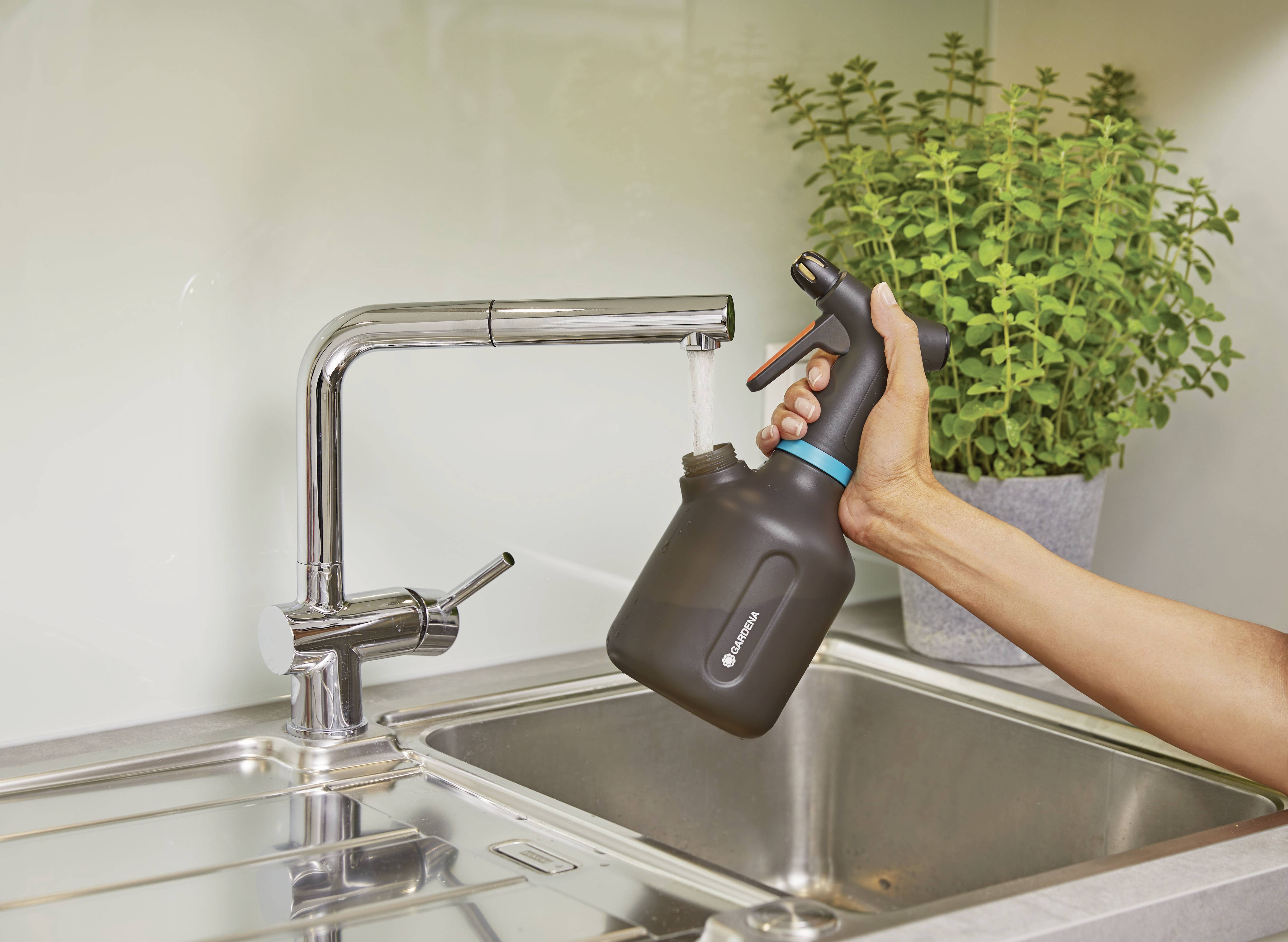 Eine Hand hält eine schwarze Sprühflasche unter einem Wasserhahn, aus dem Wasser fließt. Daneben steht ein Topf mit einer grünen Pflanze.
