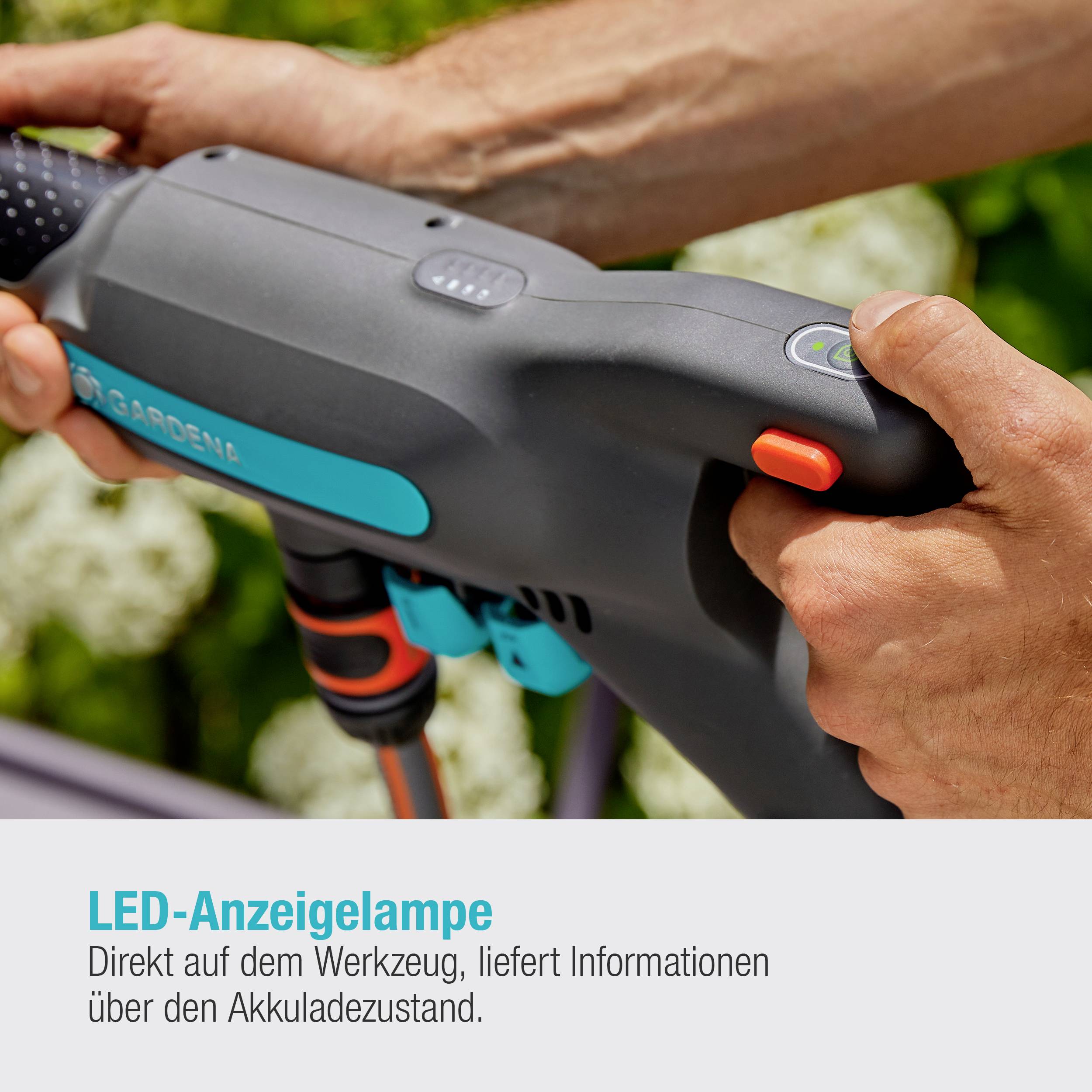 Person hält ein Gartengerät mit LED-Anzeigelampe, die den Akkuladestand zeigt. Hintergrund unscharf mit grüner Vegetation.