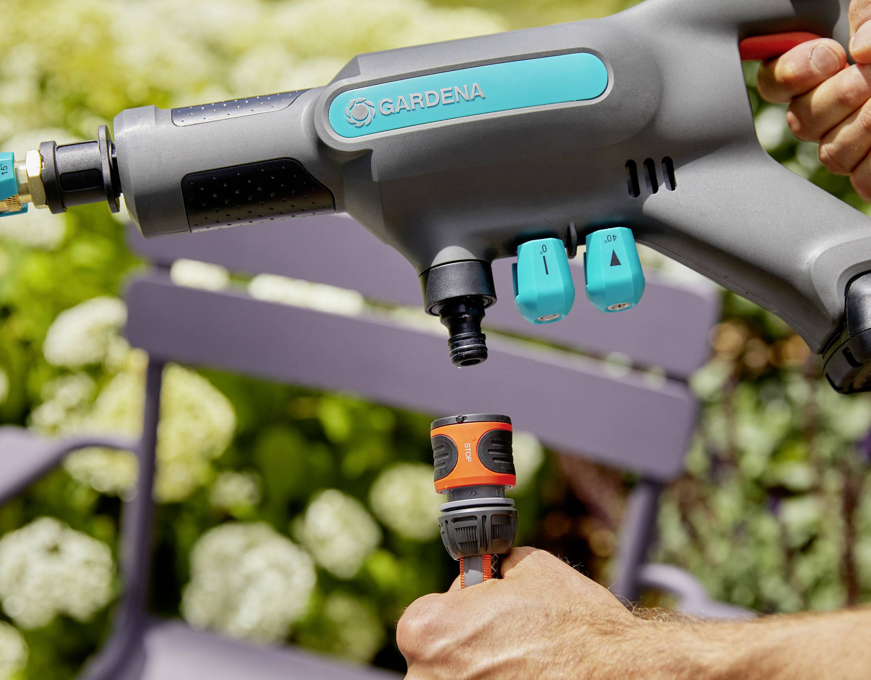 'Eine Person verbindet einen GARDENA-Wasseranschluss mit einer Sprühpistole im Garten, Hintergrund zeigt Pflanzen und Gartenstuhl.'