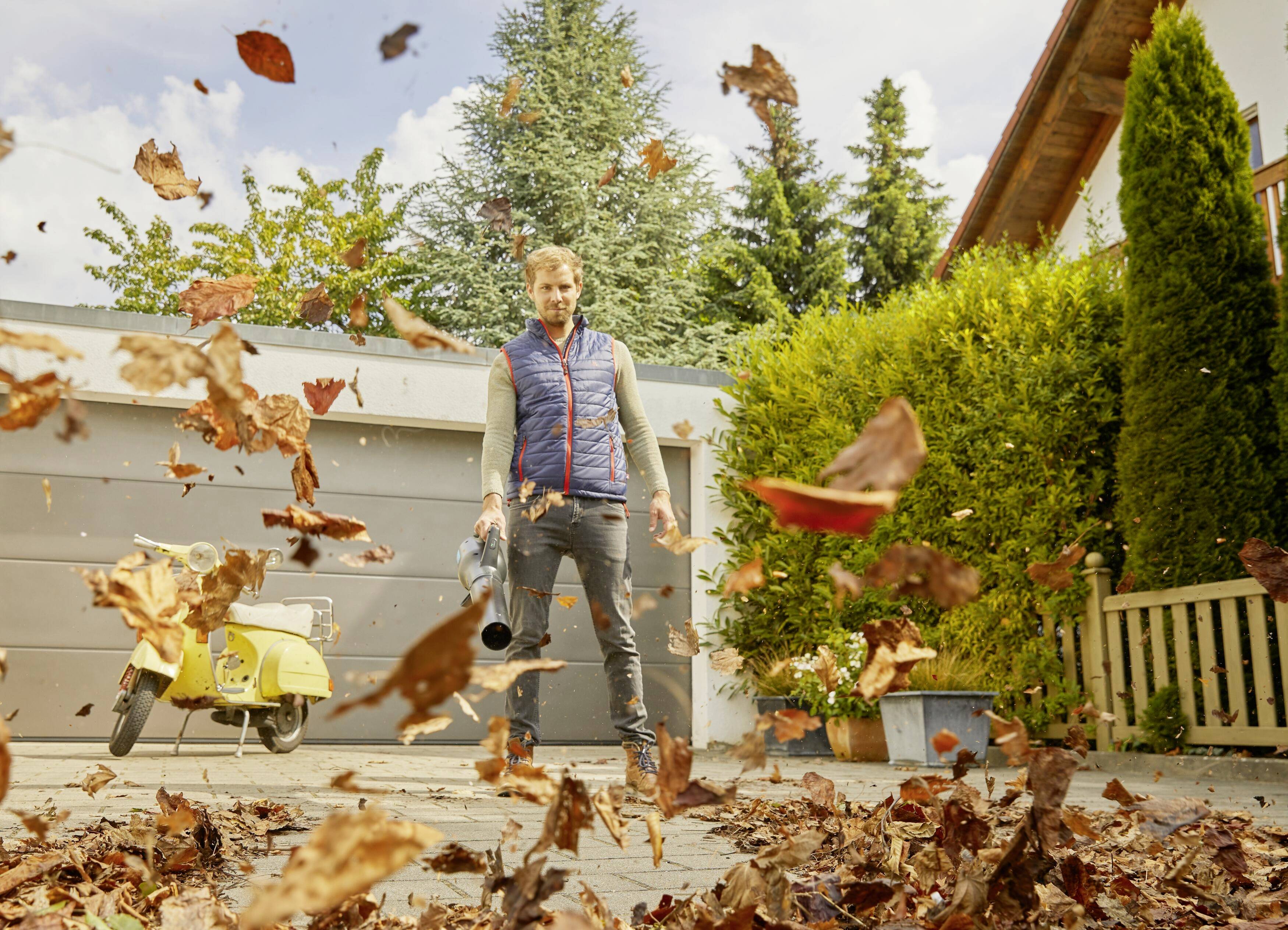 Ein Mann bläst herbstliche Blätter mit einem Laubbläser in einem Garten weg. Im Hintergrund sind Bäume und ein gelbes Motorrad zu sehen.