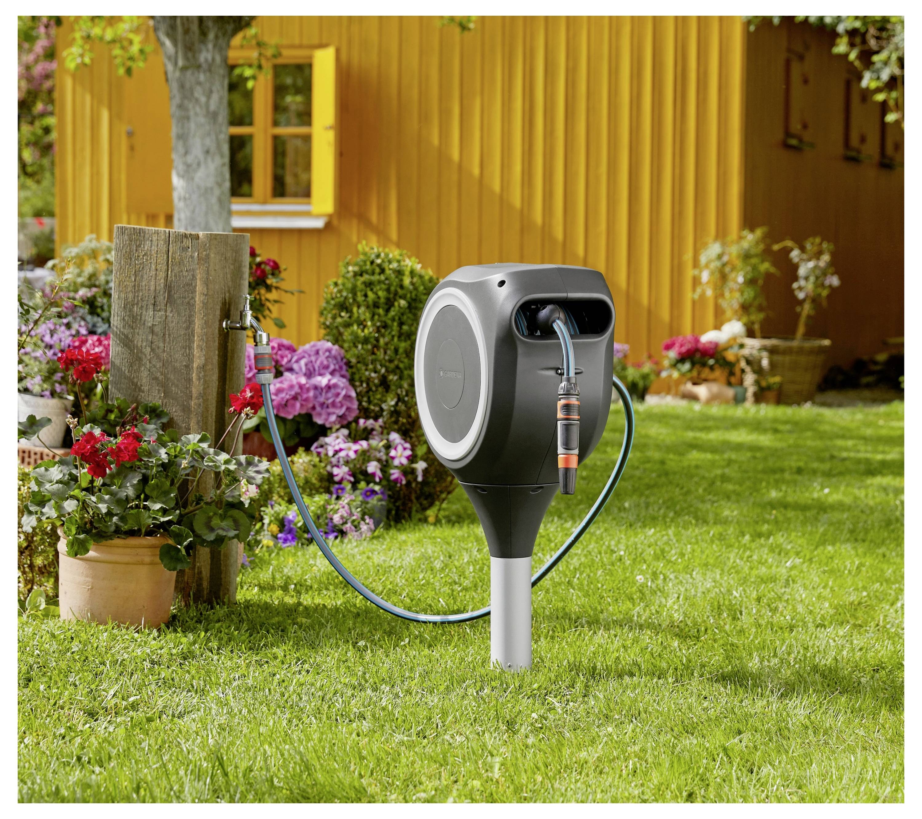 Eine moderne ausziehbare Gartenschlauchtrommel, die auf einem Pfosten in einem Blumengarten neben einem gelben Haus montiert ist.