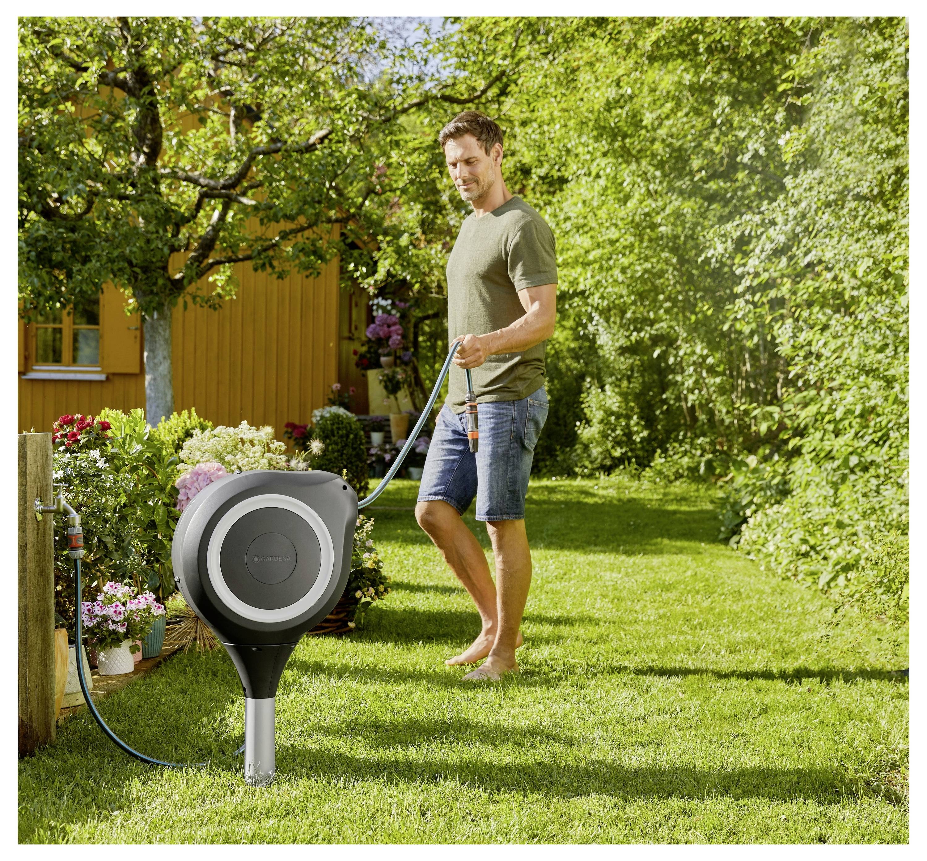 Mann, der Blumen in einem sonnigen Garten neben einer modernen Schlauchrolle und einem gelben Haus gießt.