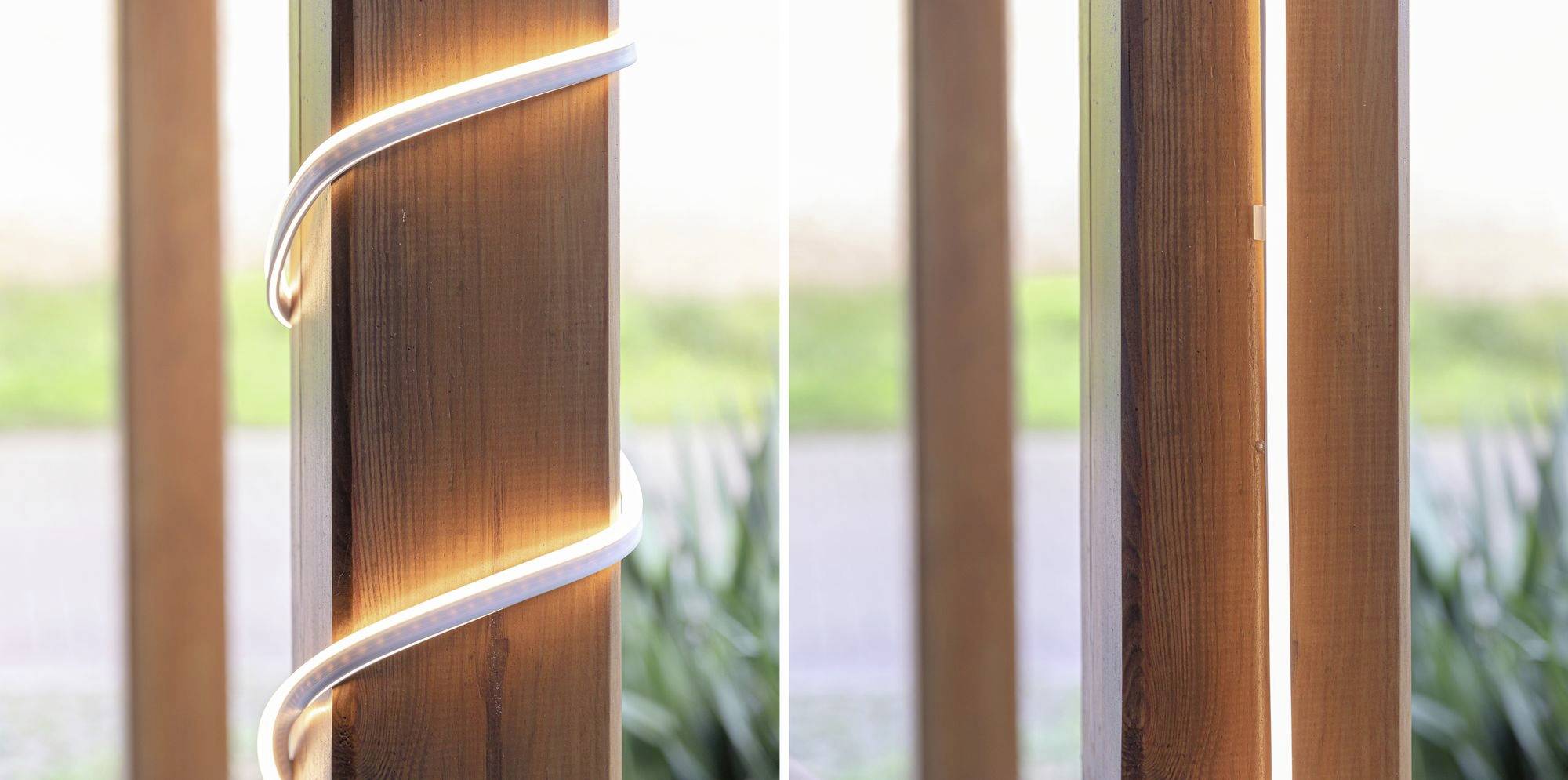 Holzpfosten mit LED-Beleuchtung: links umwickelte LED-Leiste, rechts vertikale LED-Linie. Umgeben von verschwommenem Grün.