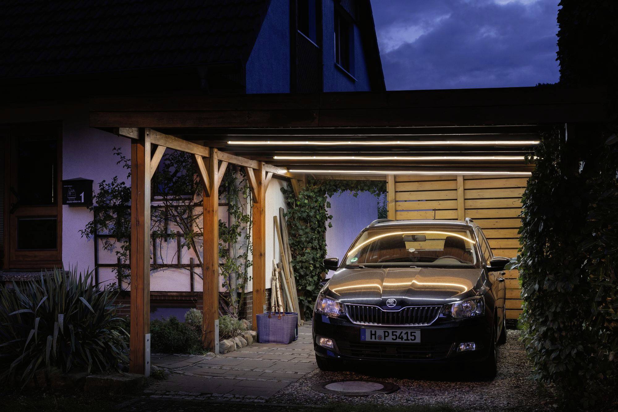 Ein Auto parkt nachts unter einem überdachten Carport neben einem Haus. Lichter am Carport leuchten den Bereich aus.