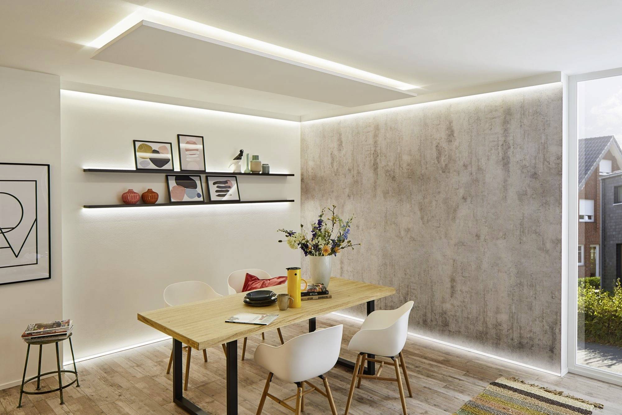 Esszimmer mit Holztisch und weißen Stühlen, dekoriert mit Blumen in einer Vase. Moderne Beleuchtung und Bilder an der Wand.