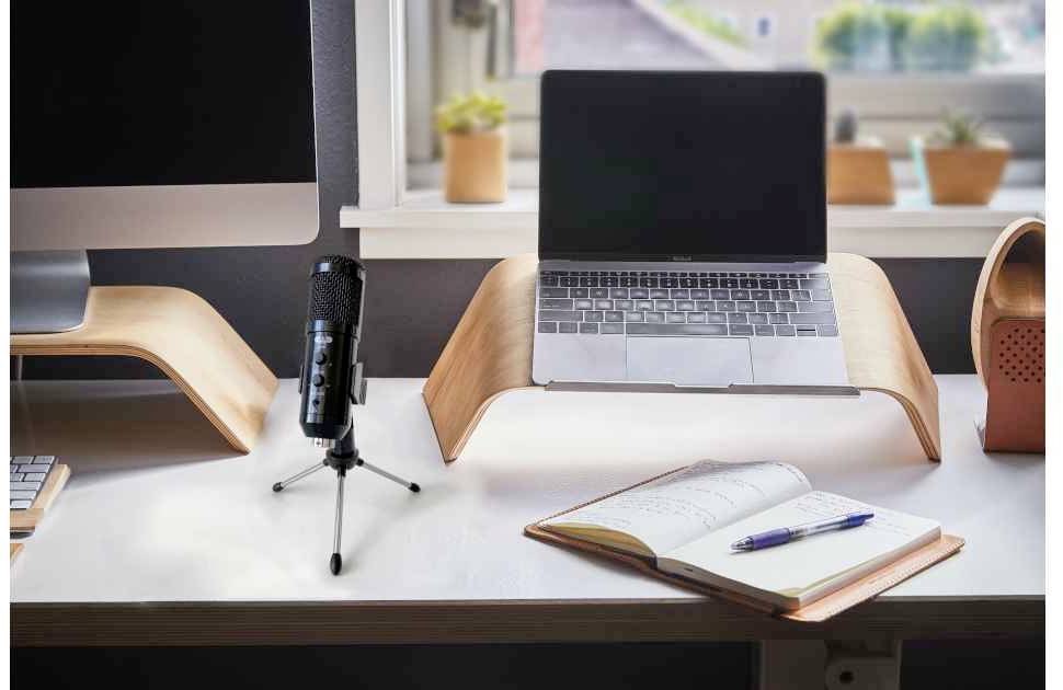 Schreibtisch mit offenem Laptop auf Holzständer, Notizbuch, Stift und Mikrofon; Fenster im Hintergrund mit Pflanzen.