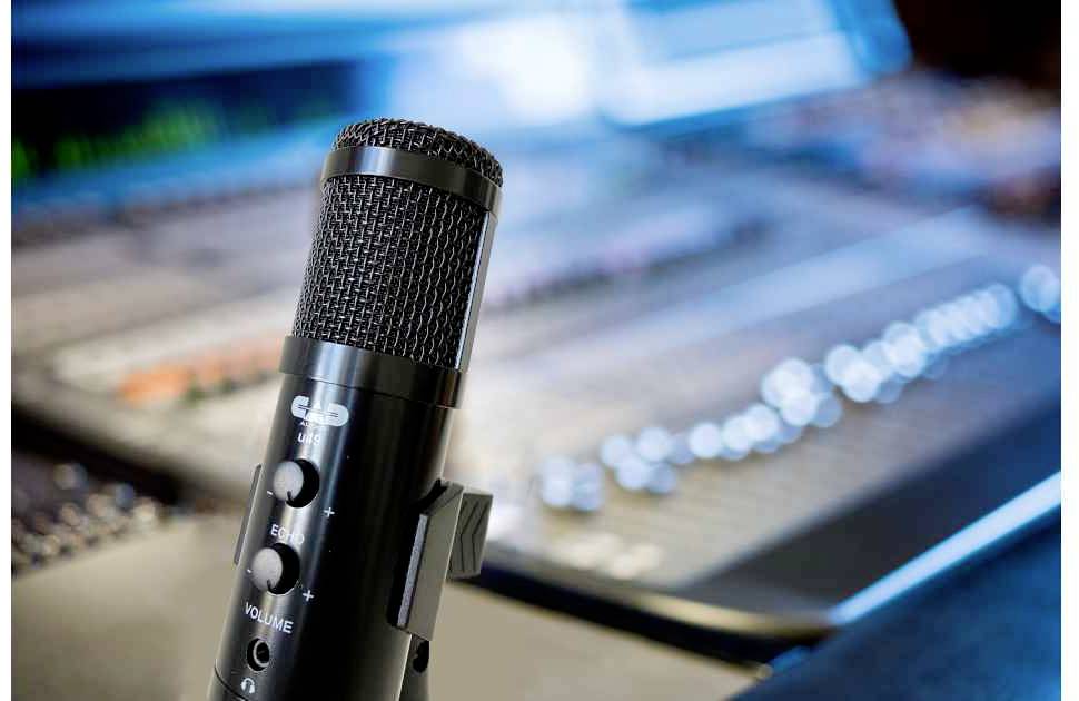 Ein Mikrofon steht im Vordergrund vor einem unscharfen Mischpult in einem Tonstudio, was auf professionelle Audioaufnahmen hinweist.