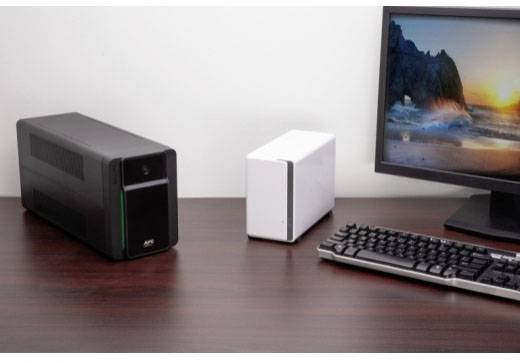 'Computertisch mit Tastatur, Monitor, schwarzem UPS-Gerät links und weißem Speichergerät in der Mitte. Bildschirm zeigt Strandbild.'