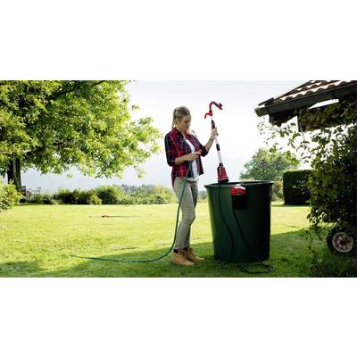 Eine Frau steht im Garten neben einem großen Wassertank und hält eine Pumpe. Sie legt den Schlauch in den Tank, um Wasser zu fördern.