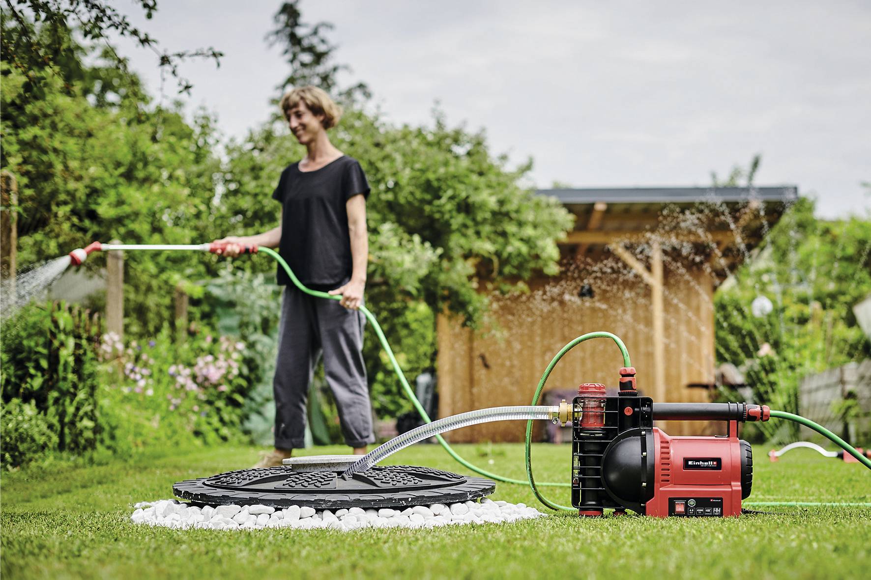 Eine Person gießt einen Garten mit einem Schlauch, der an eine rote elektrische Wasserpumpe angeschlossen ist. Im Hintergrund steht ein Holzschuppen.