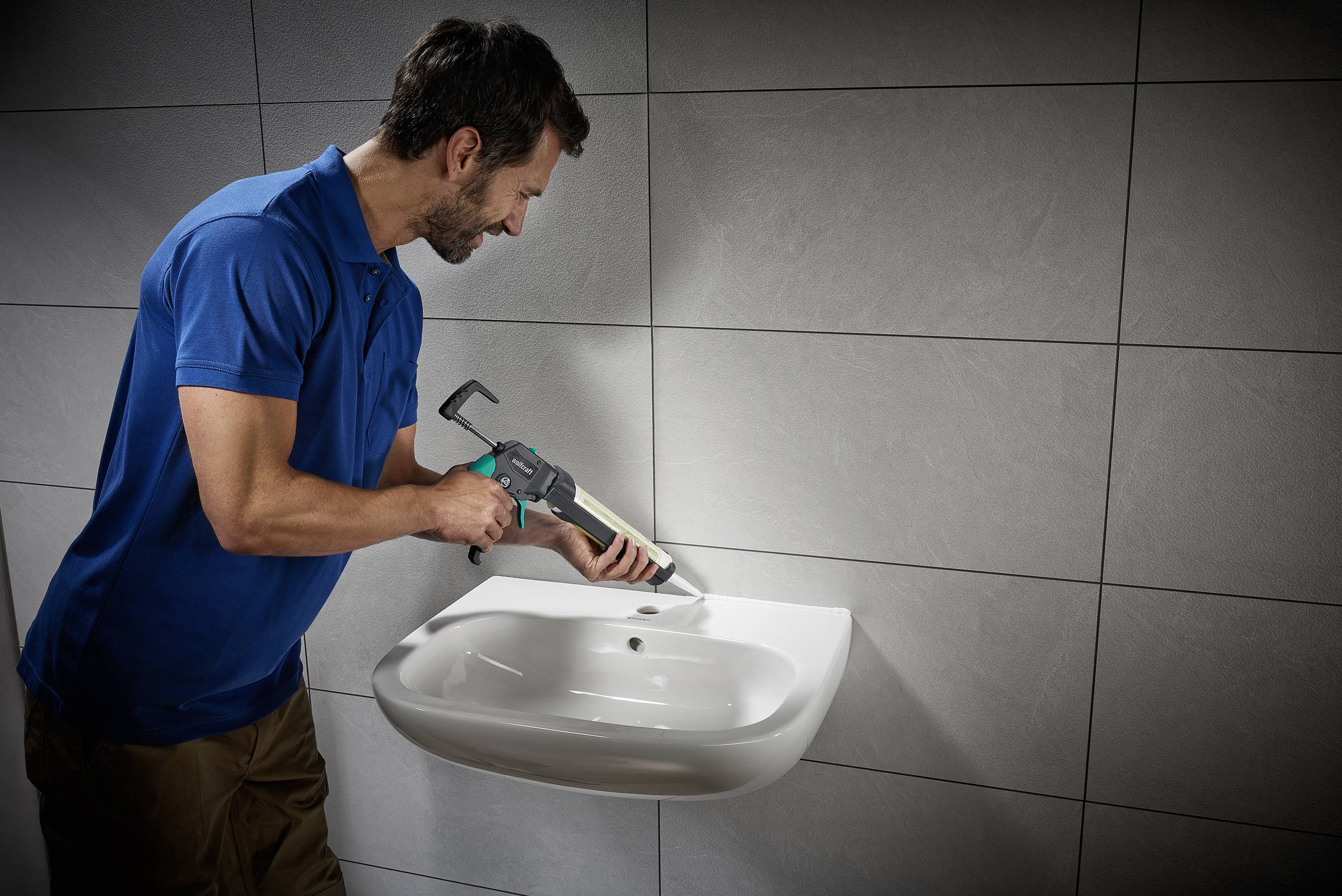 Ein Mann in blauem Hemd dämmt mit einem Dichtstoffpistole die Ränder eines an der Wand montierten Waschbeckens in einem Badezimmer ab.