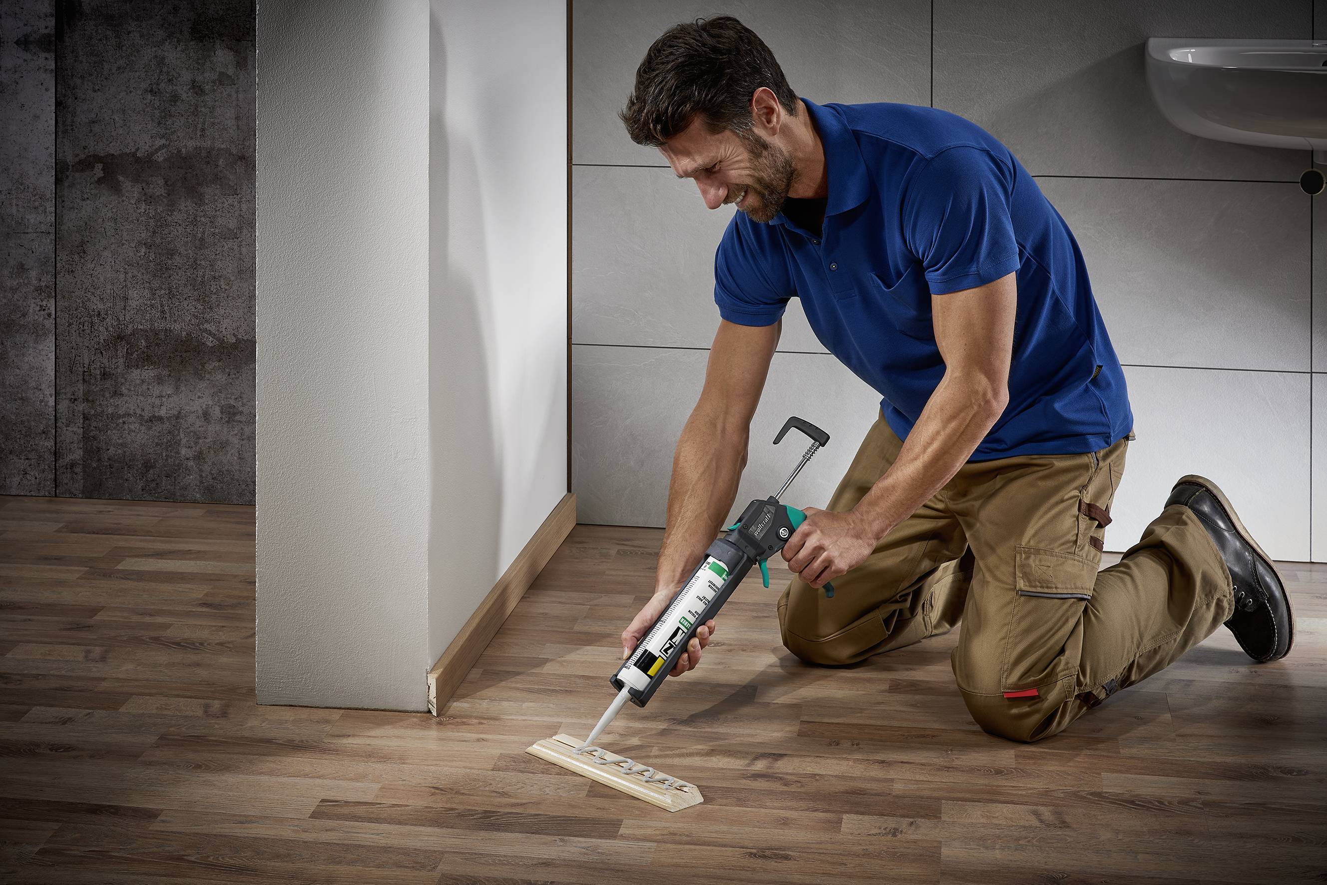 Ein Mann in Arbeitskleidung verwendet eine Klebepistole, um ein Holzstück auf einem Holzboden zu befestigen.