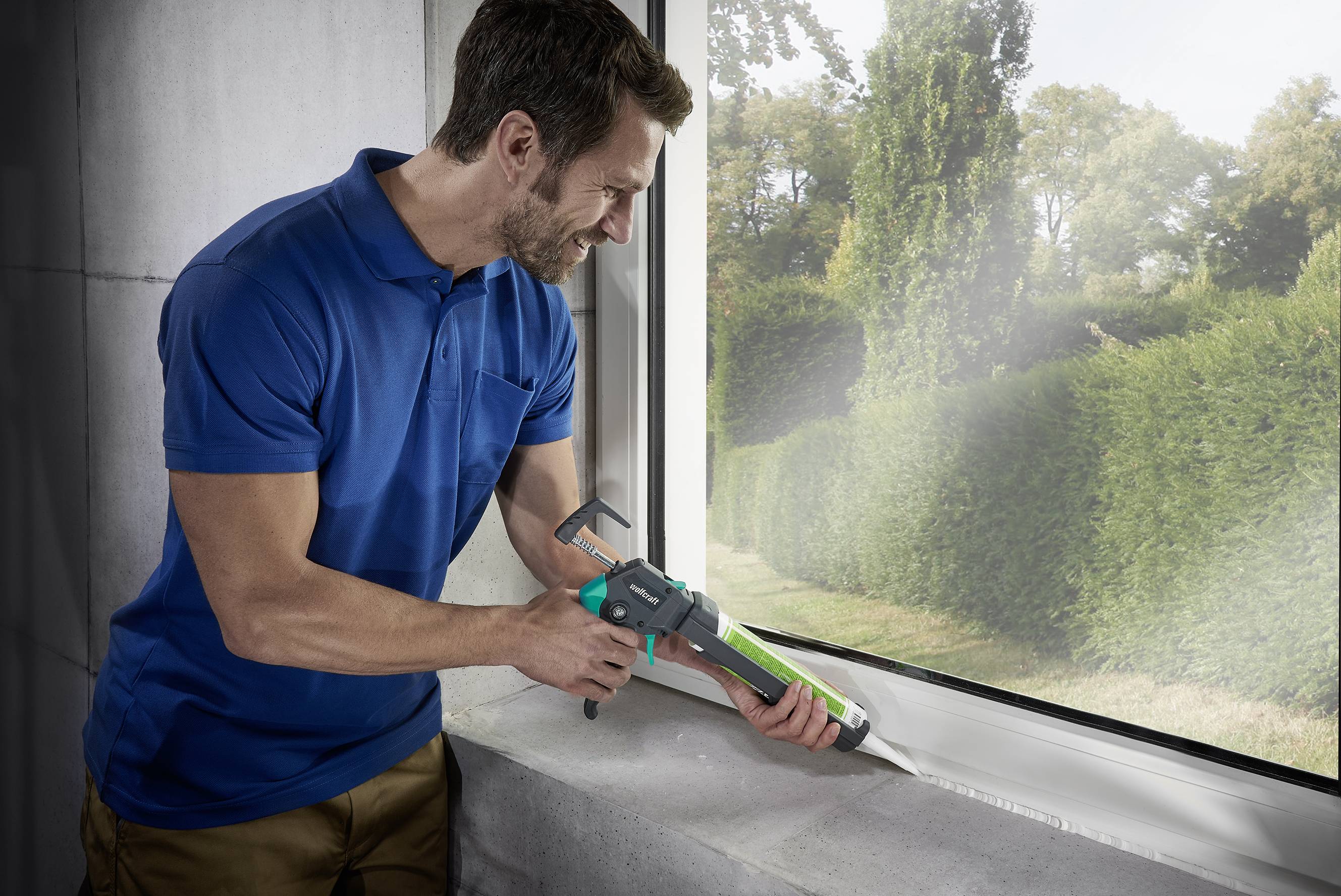 Ein Mann trägt ein blaues Poloshirt und versiegelt ein Fenster mit einer Kartuschenpistole in einem sonnigen Raum mit Blick auf Gartenhecken.