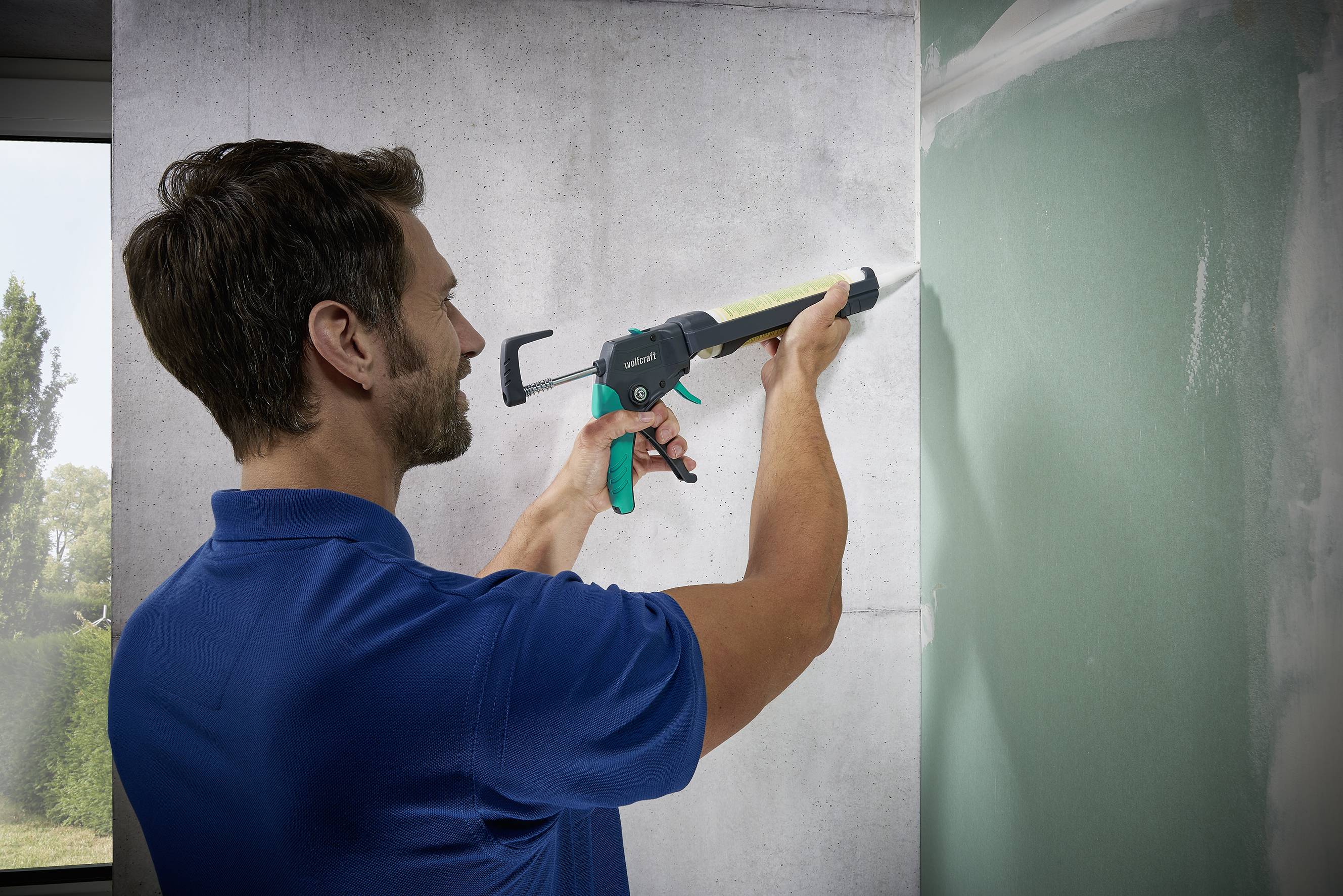 Ein Mann im blauen Hemd verwendet eine Kartuschenpistole, um Dichtmasse auf eine Wand aufzutragen. Links ist ein Fenster mit Bäumen zu sehen.