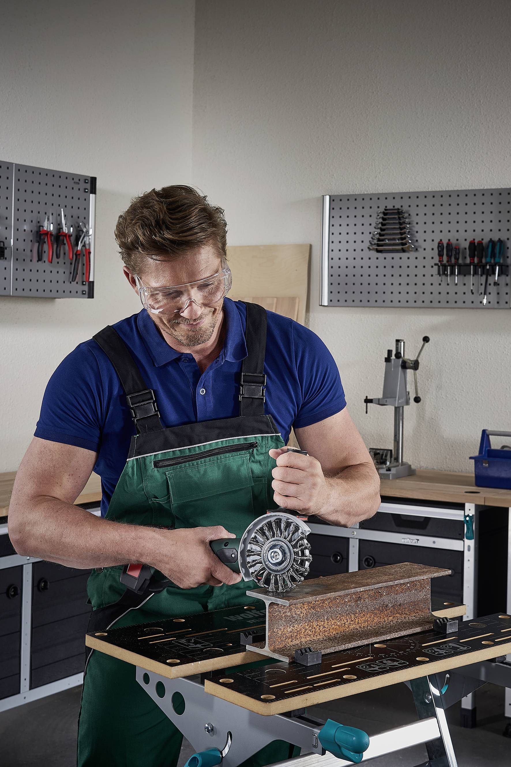 Ein Mann in Arbeitskleidung schleift mit einem Winkelschleifer Metall in einer Werkstatt. Im Hintergrund sind Werkzeuge an der Wand.
