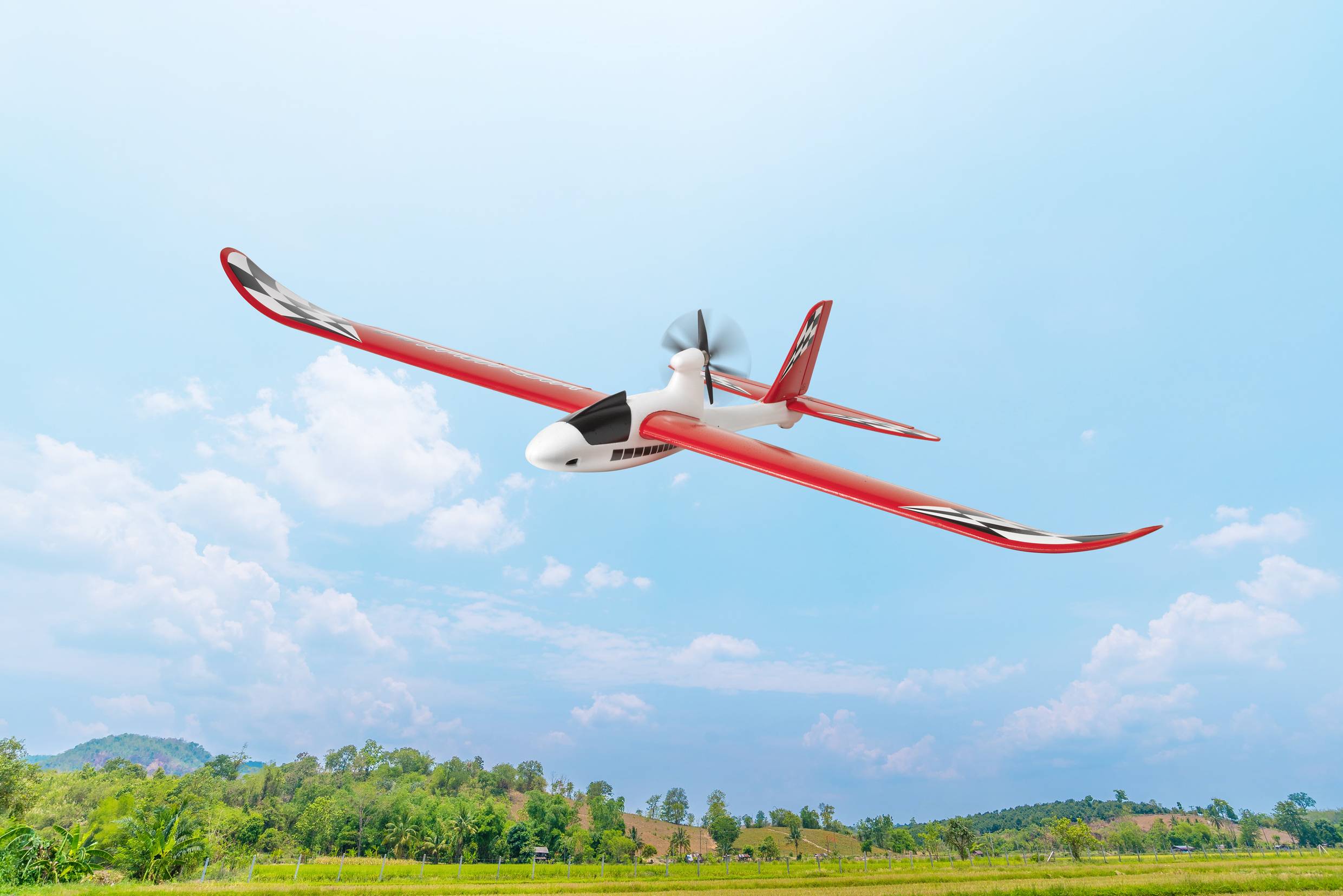 Eine rote Drohne fliegt am klaren Himmel über einer grünen Landschaft mit Bäumen und Hügeln im Hintergrund.