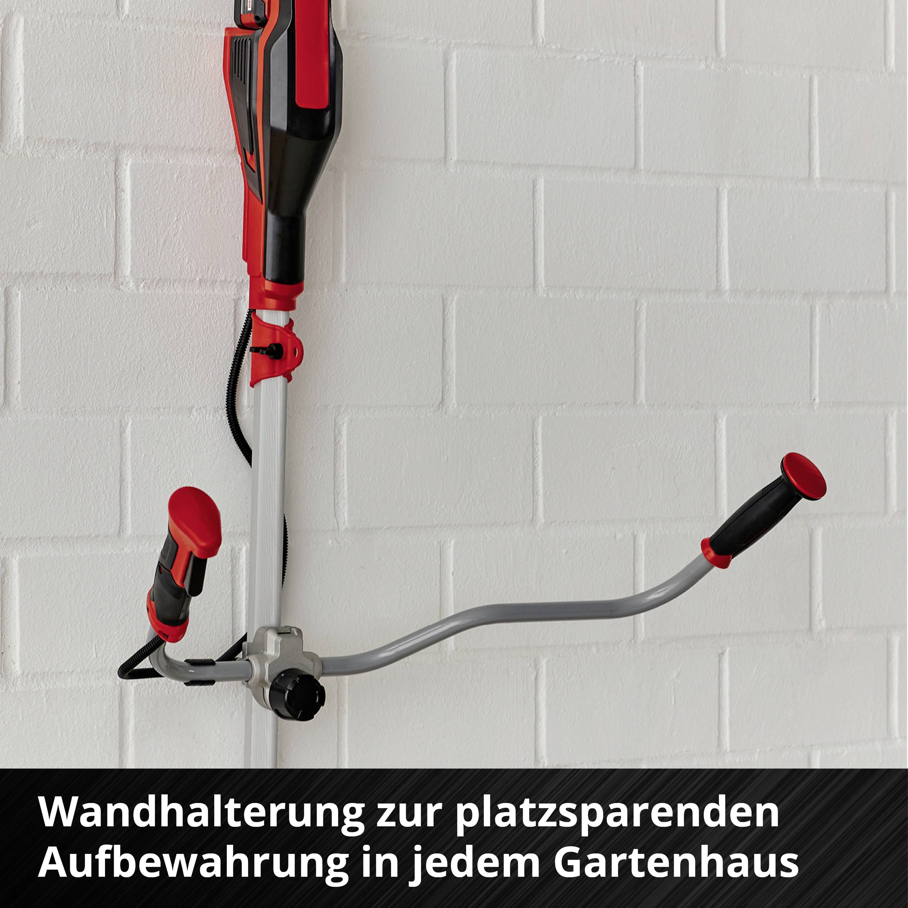 Ein roter Rasentrimmer hängt an einer Wandhalterung vor einer weißen Ziegelwand. Darunter steht: 'Wandhalterung zur platzsparenden Aufbewahrung in jedem Gartenhaus'.