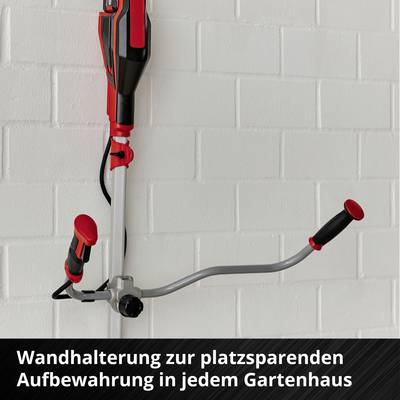 Ein roter Rasentrimmer hängt an einer Wandhalterung vor einer weißen Ziegelwand. Darunter steht: 'Wandhalterung zur platzsparenden Aufbewahrung in jedem Gartenhaus'.