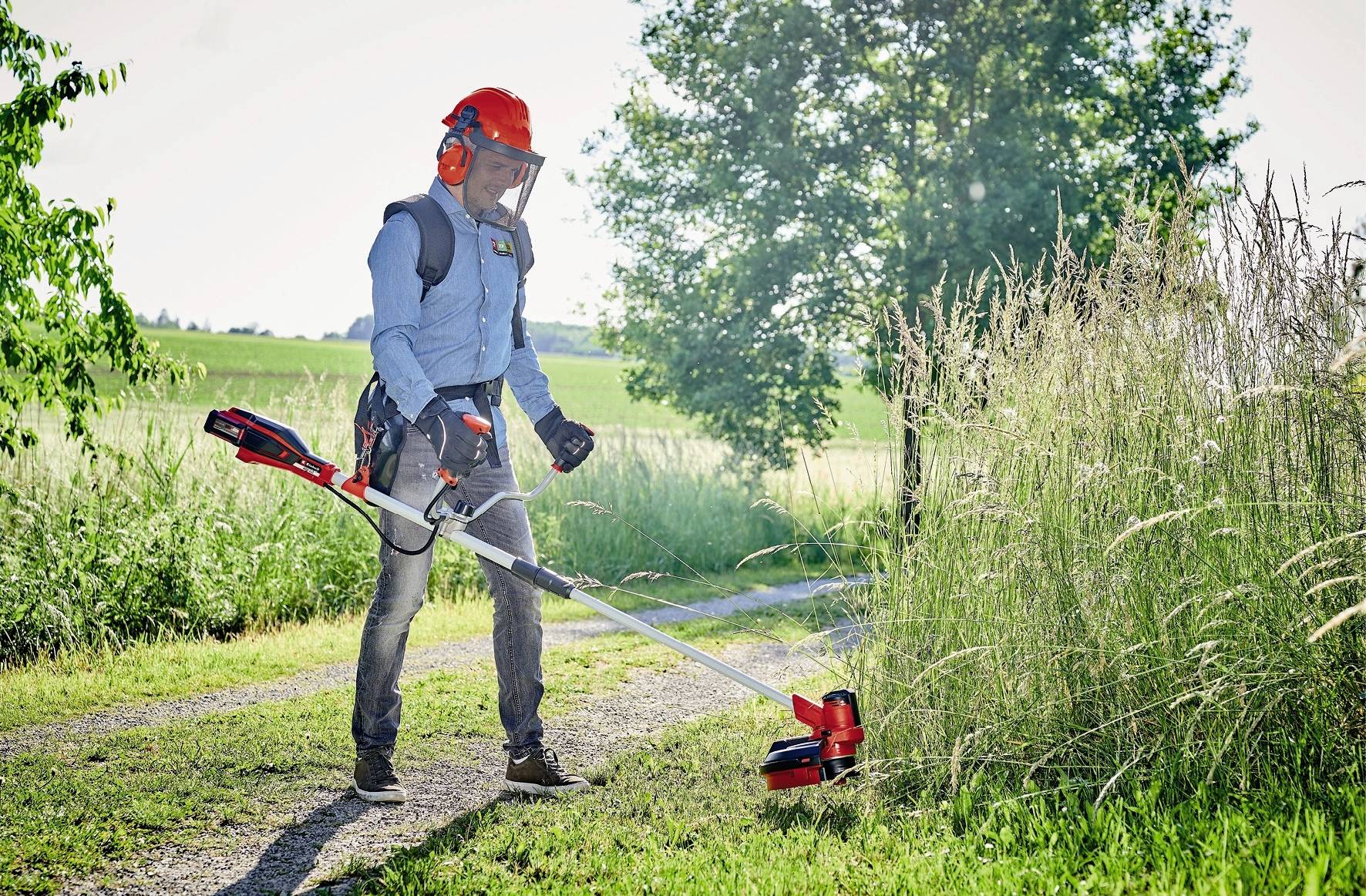 Eine Person mit Schutzkleidung nutzt einen Rasentrimmer, um hohes Gras neben einem Weg in einer ländlichen Umgebung zu schneiden.