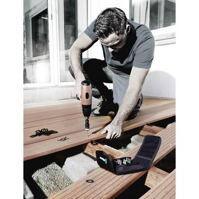 Mann baut eine Holzterrasse, kniend mit einem Elektrowerkzeug. Werkzeugtasche mit Bits im Vordergrund. Hausfassade im Hintergrund.