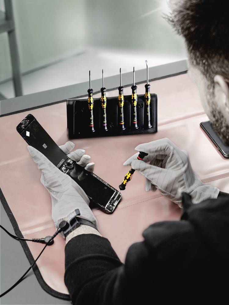 Eine Person repariert ein Smartphone auf einer Werkbank, hält das Gerät mit einer Pinzette, umgeben von Mikro-Werkzeugen.