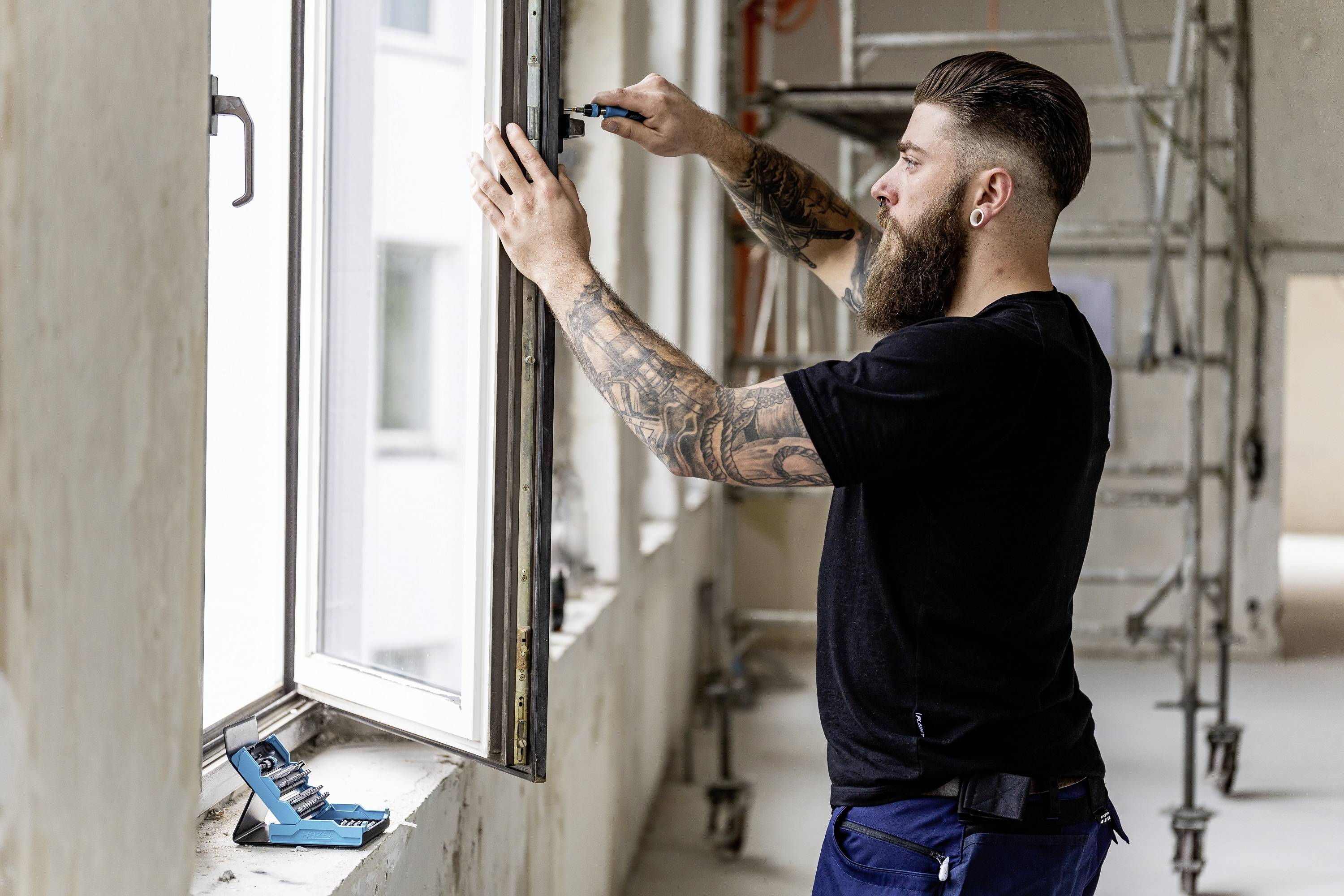 Ein Mann mit Bart installiert ein Fensterrahmen in einem Gebäude. Im Hintergrund ist ein Gerüst erkennbar.