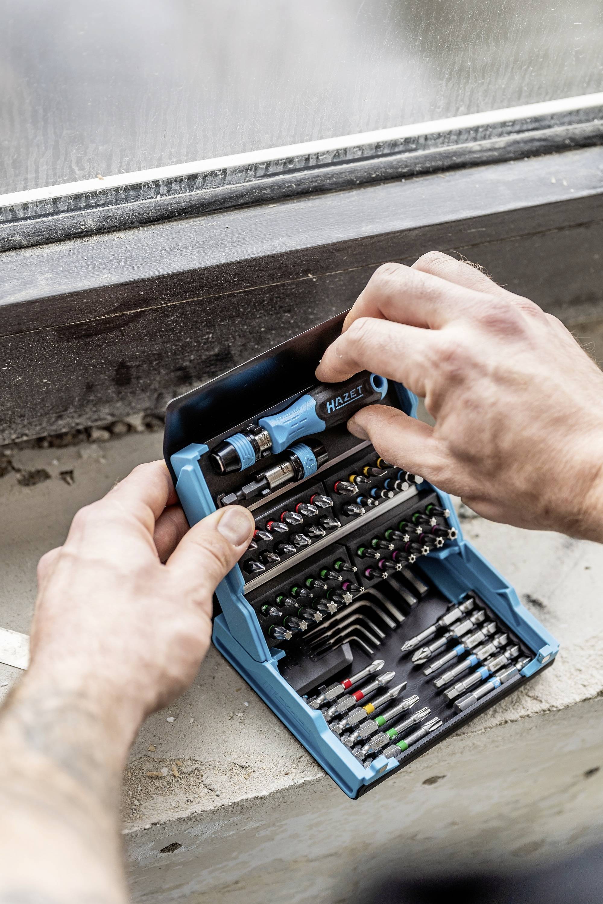 Person öffnet ein blaues Werkzeugset mit verschiedenen Aufsätzen und Bits auf einer Fensterbank.