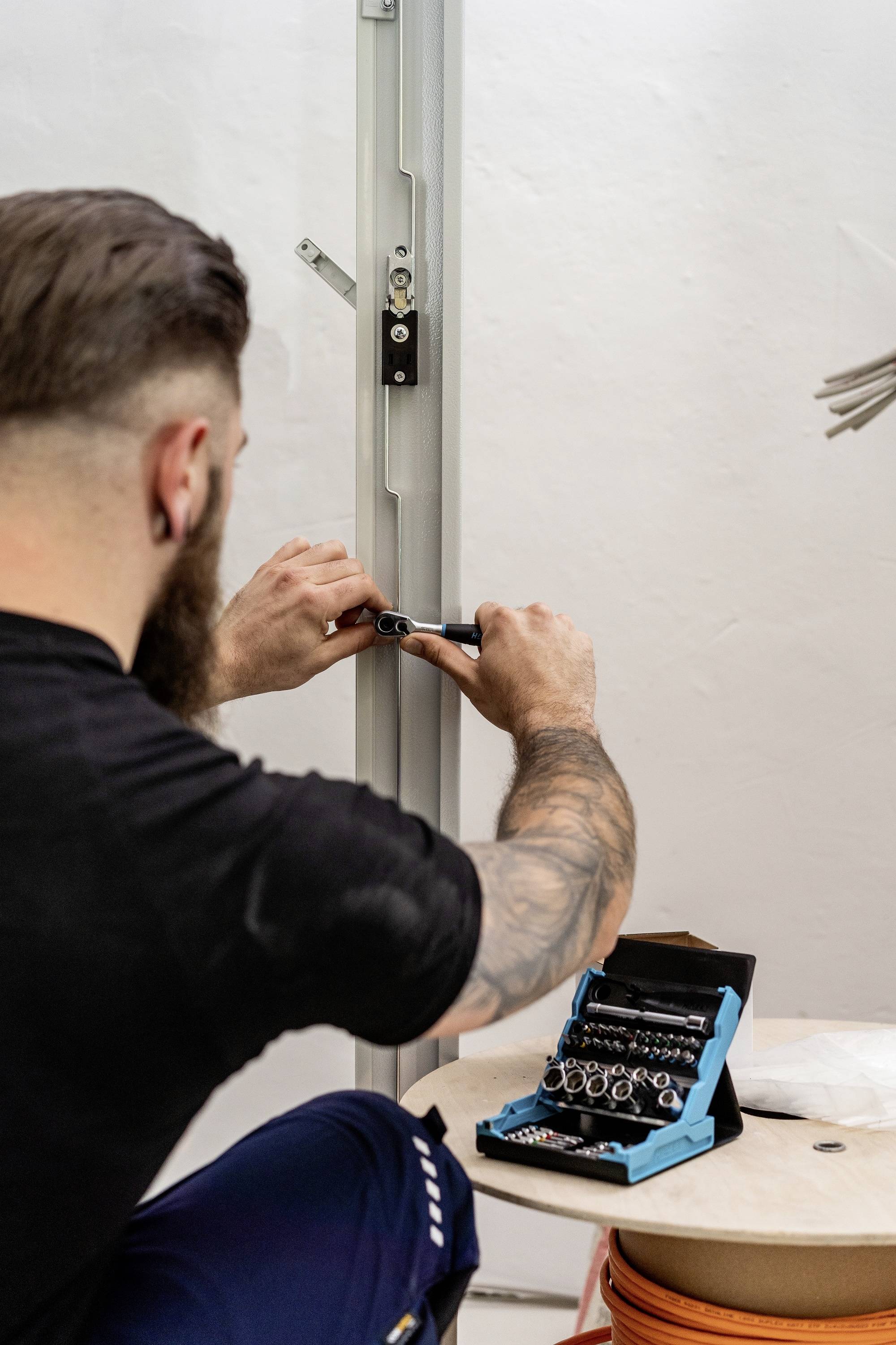 Eine Person arbeitet an einem metallischen Rahmen mit Werkzeugen. Ein offener Werkzeugkasten mit Schrauben liegt auf einem Tisch in der Nähe.