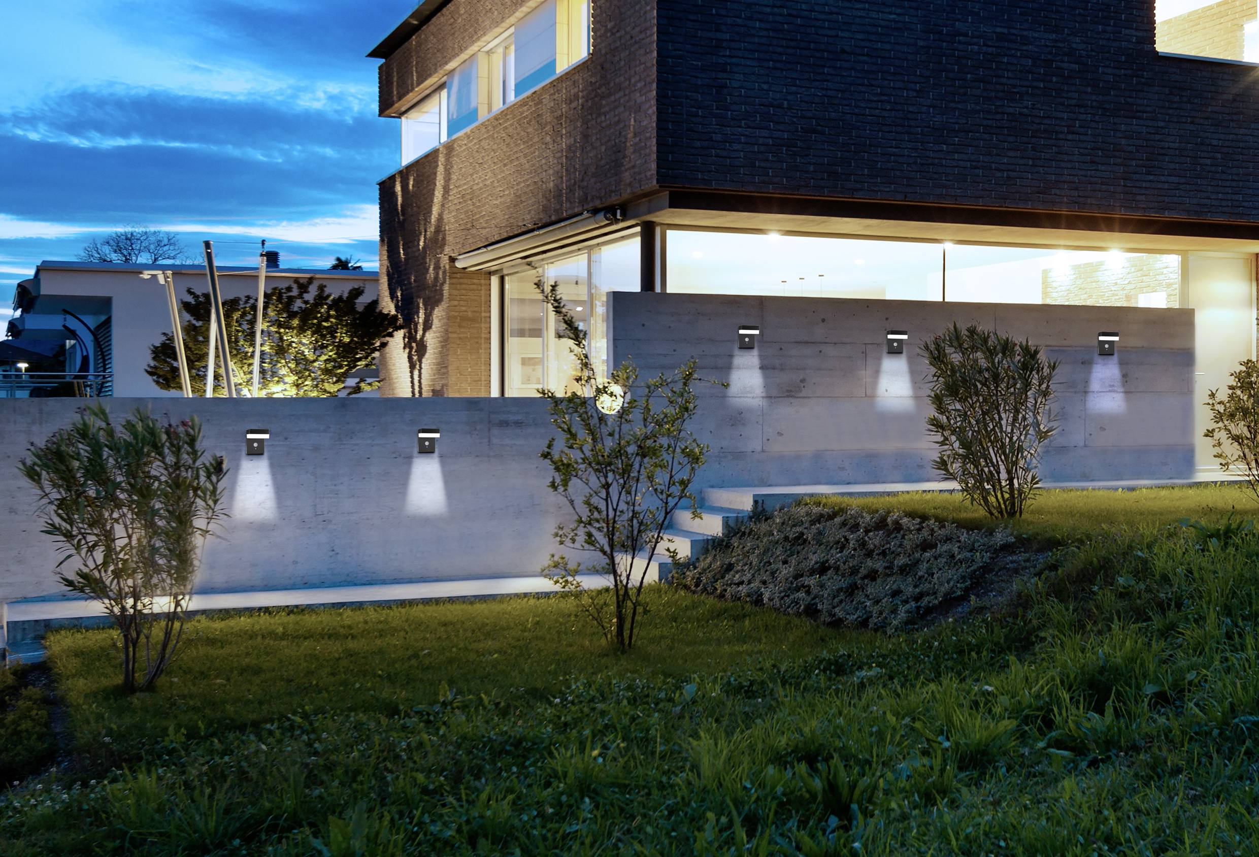 Ein modernes Haus bei Dämmerung mit leuchtender Außenbeleuchtung auf der Betonwand. Schritte führen durch den gepflegten Garten.