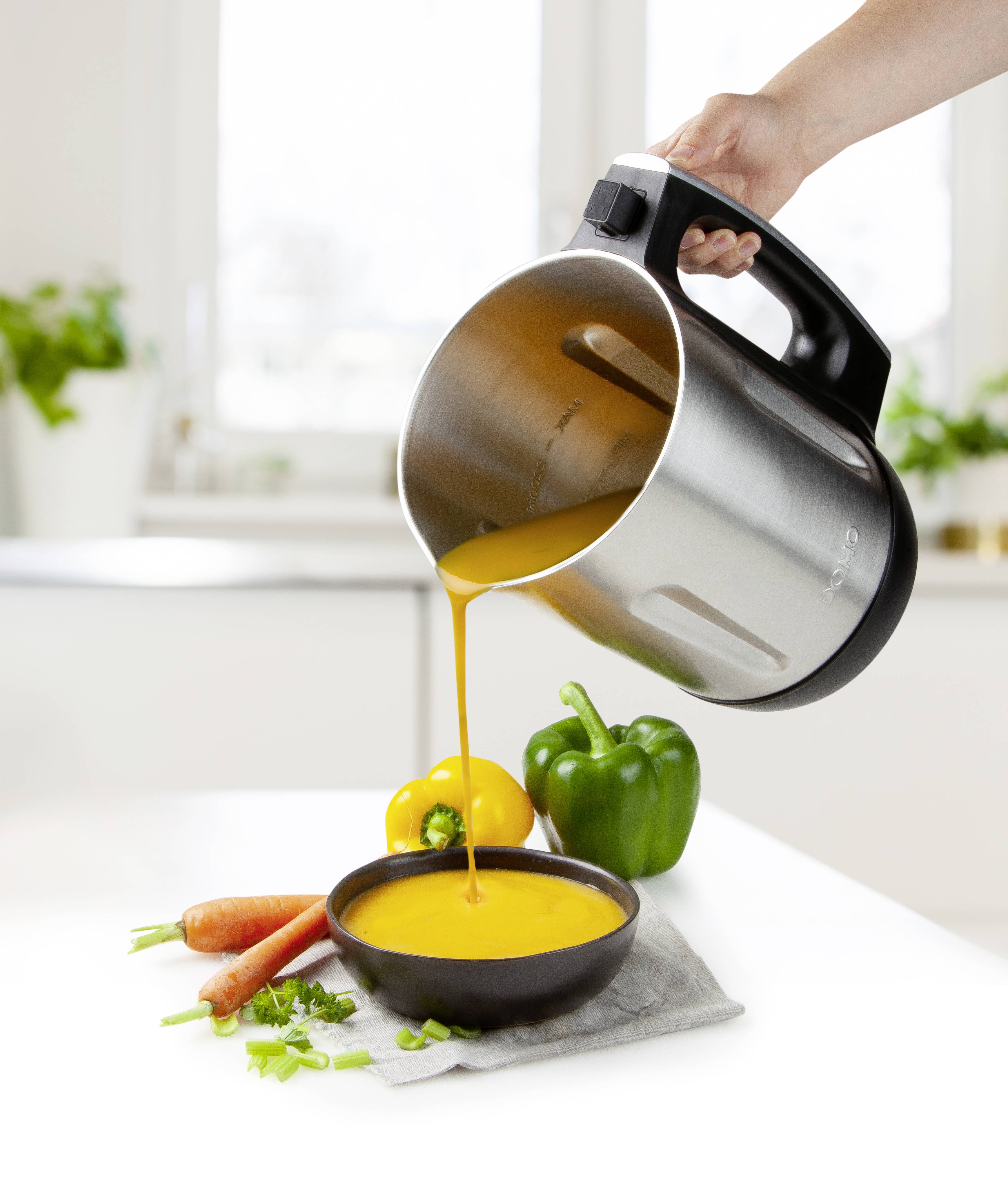 Eine Hand gießt gelbe Suppe aus einem Mixer in eine Schale auf einem Tisch. Daneben liegen Paprika, Karotten und Kräuter.