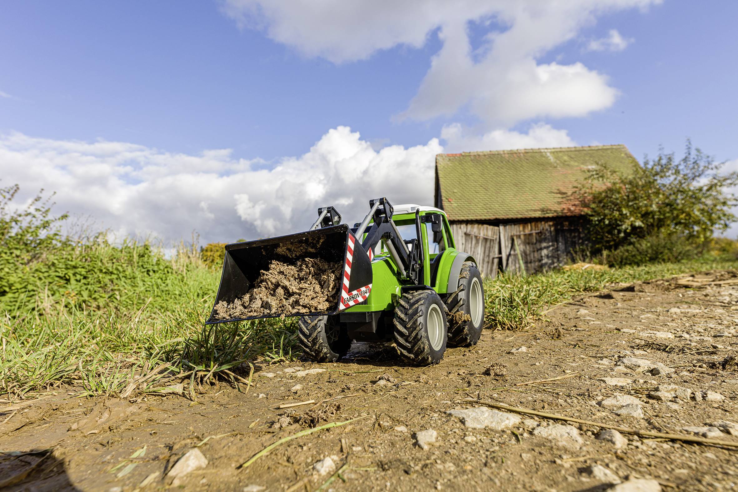 Ein grüner Traktor mit Frontlader transportiert Erde auf einem ländlichen Feldweg, im Hintergrund steht eine alte Scheune.