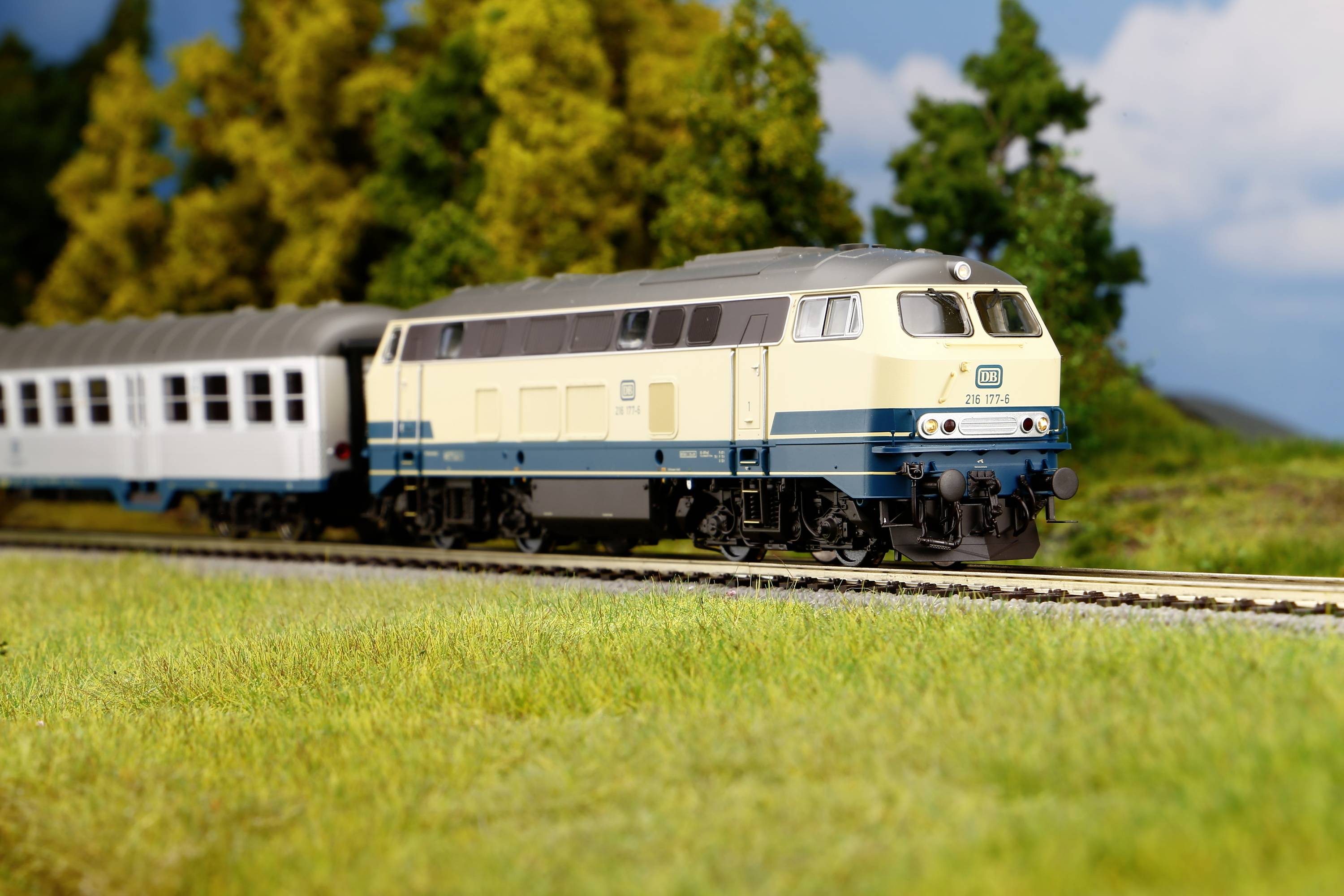 Ein Modellzug fährt durch eine ländliche Szenerie mit Bäumen im Hintergrund unter blauem Himmel. Lokomotive mit beigen und blauen Farben.