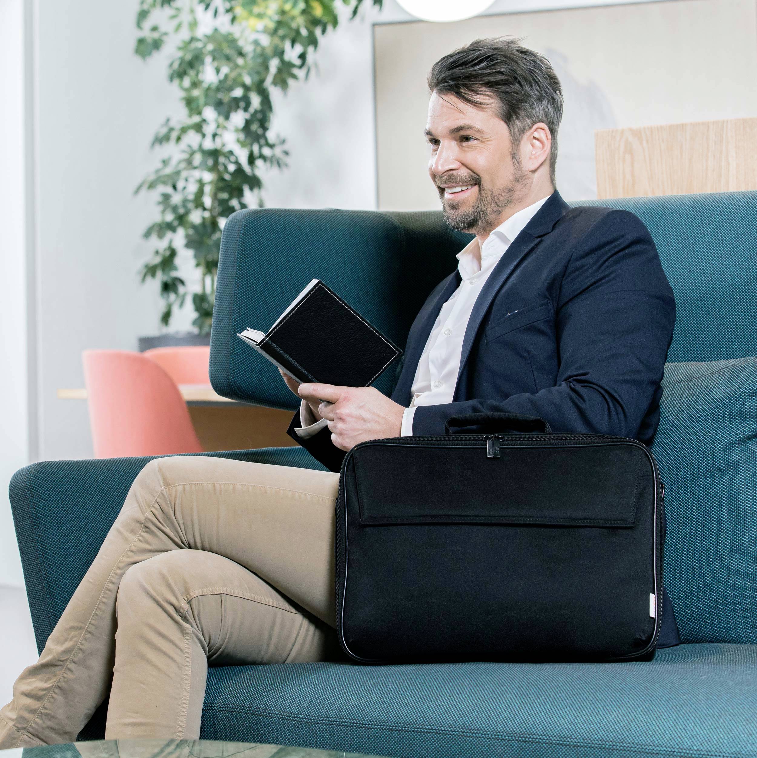 Ein Mann in einem Anzug sitzt auf einem Sofa und hält ein Tablet. Im Hintergrund sind Pflanzen und ein heller Raum zu sehen.