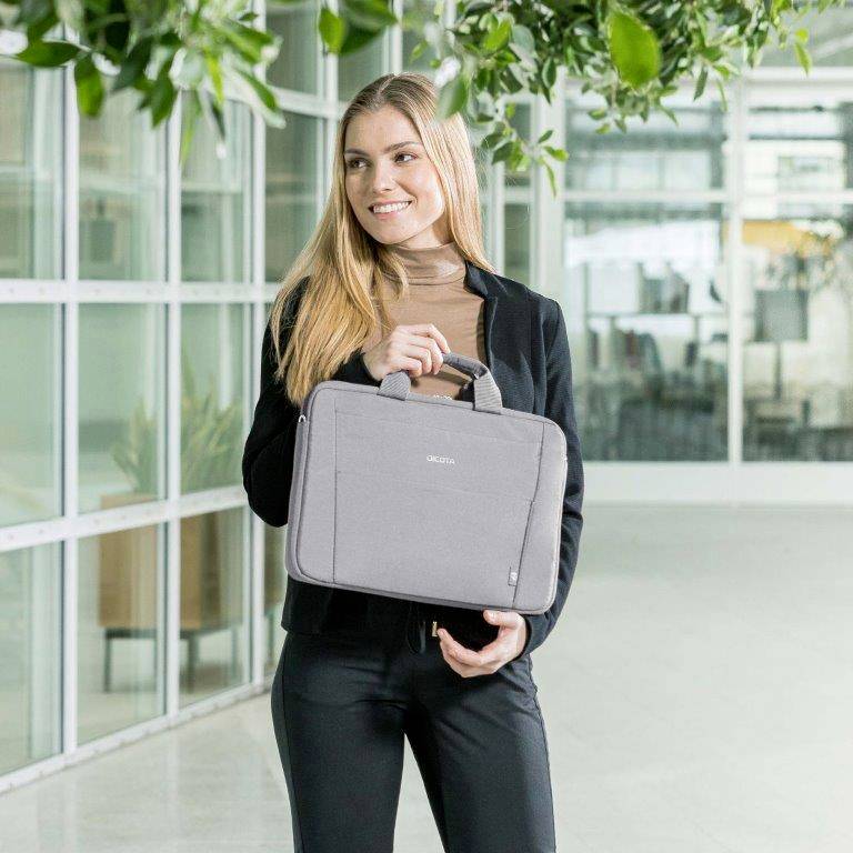 Eine lächelnde Frau steht in einem modernen Bürogebäude und hält eine graue Laptop-Tasche in der Hand. Pflanzen hängen von der Decke.