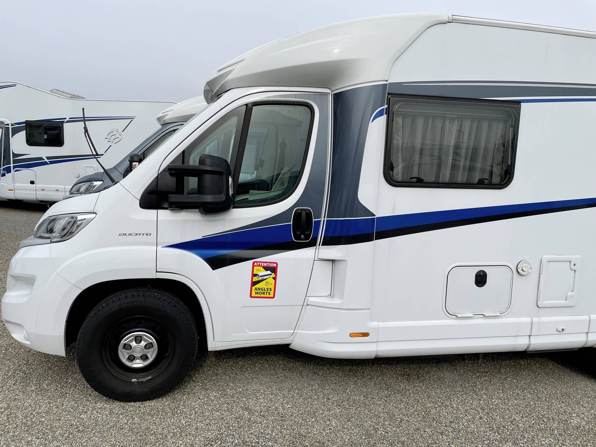 Weißes Wohnmobil mit blauem Streifen, geparkt auf einem Kiesplatz. Seitlich ein Aufkleber mit dem ADAC Testsieger-Logo.