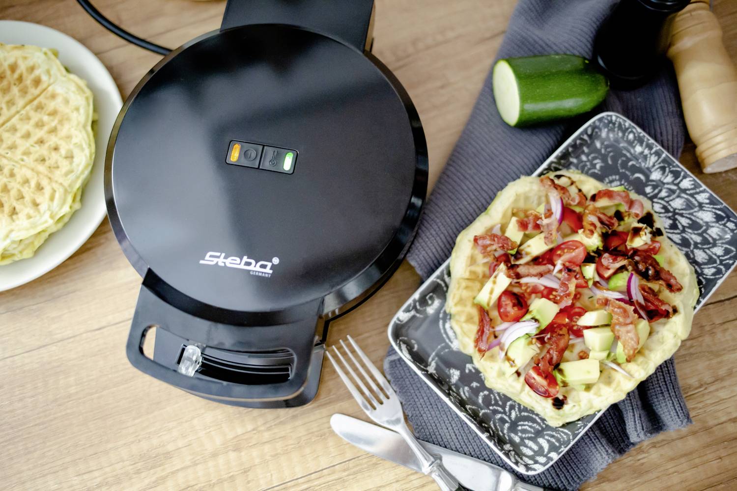 Ein Waffeleisen neben einem Teller mit einer herzhaft belegten Waffel, garniert mit Tomaten, Zucchini und Zwiebeln. Im Hintergrund weitere Waffeln.