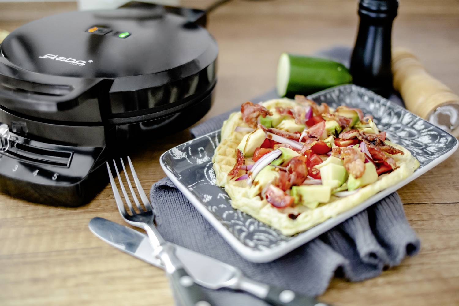 Ein Waffeleisen neben einem Teller mit herzhaften Waffeln, belegt mit frischem Gemüse und Speck. Daneben liegen Besteck und eine Salatgurke.