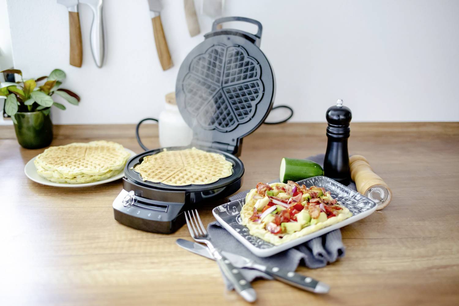 Waffeleisen auf Holztisch, öffnet sich mit zwei frisch gebackenen Waffeln. Teller daneben mit Waffel, Gemüse und Besteck.
