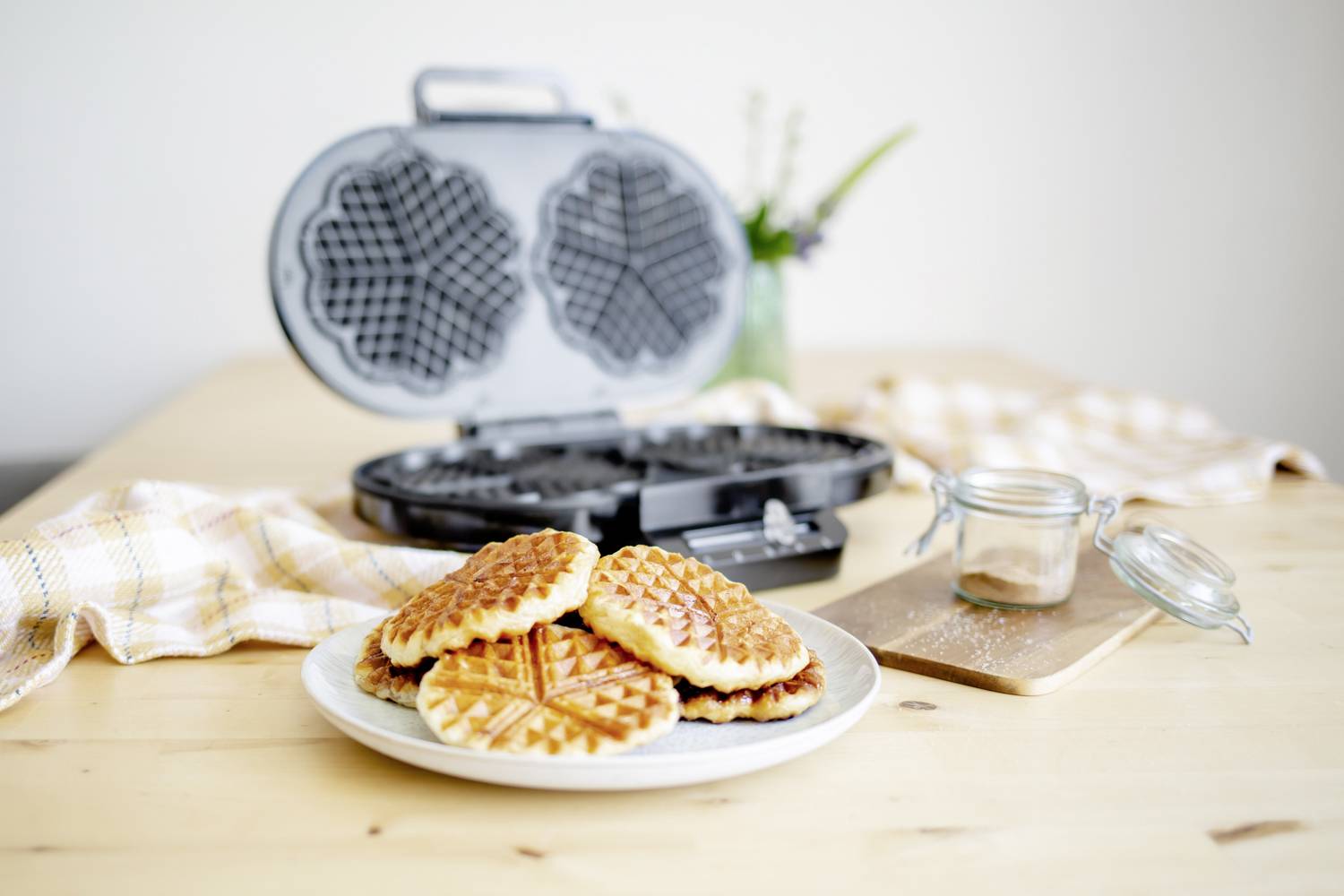 Ein Teller mit sechs Waffeln steht auf einem Holztisch, neben einem offenen Waffeleisen. Im Hintergrund Glas mit Blumen.