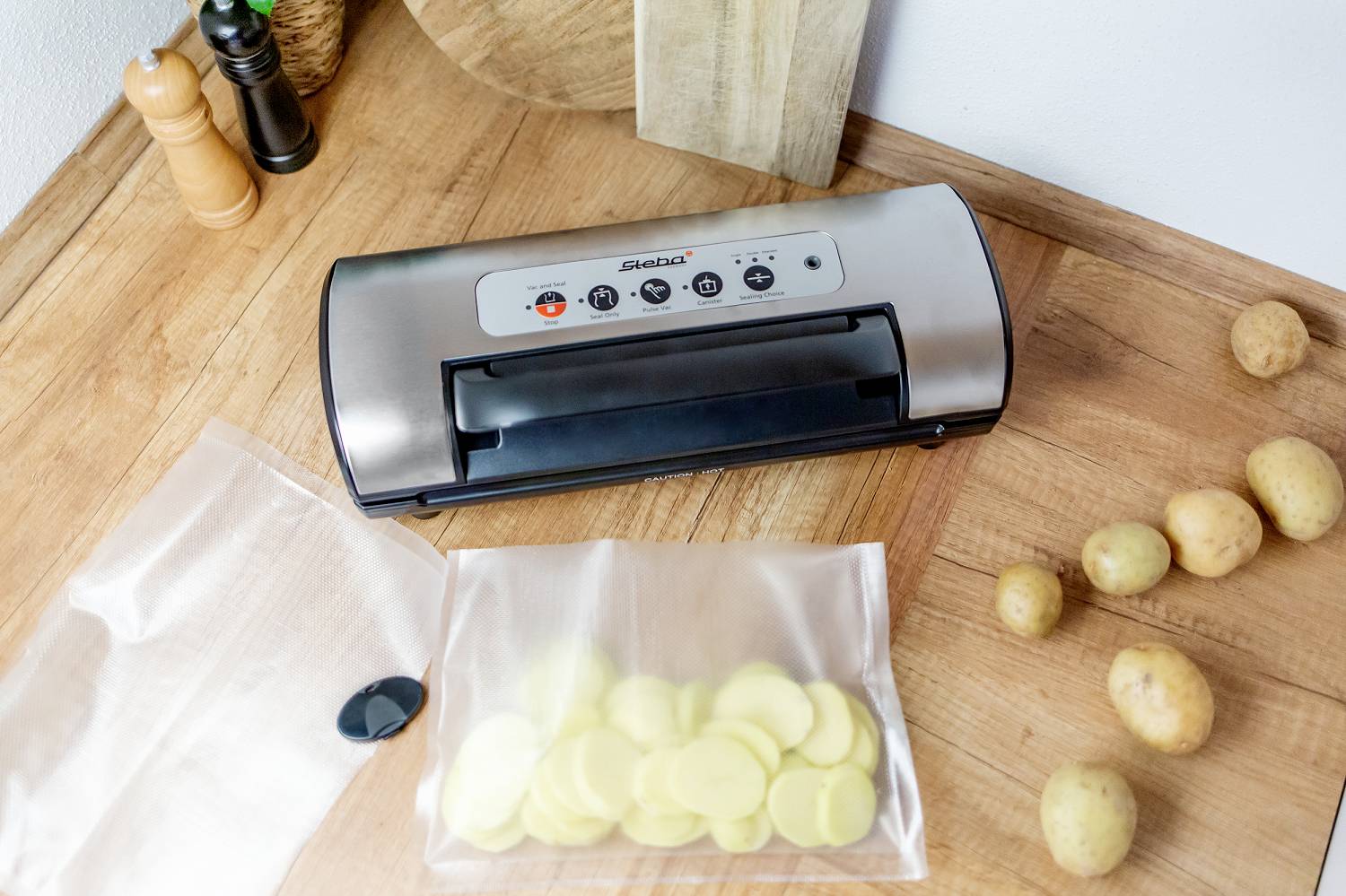 Vakuumiergerät auf Küchentheke, umgeben von Kartoffeln. Geöffneter Beutel zeigt geschnittene Kartoffelscheiben.