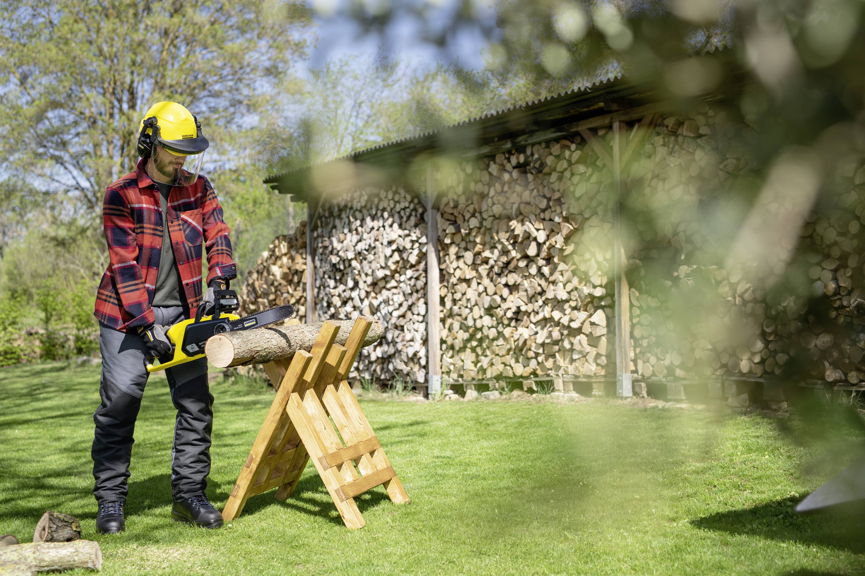 Eine Person mit Schutzhelm und Schutzbrille sägt einen Holzstamm mit einer Motorsäge im Garten. Im Hintergrund ist ein Holzlager zu sehen.