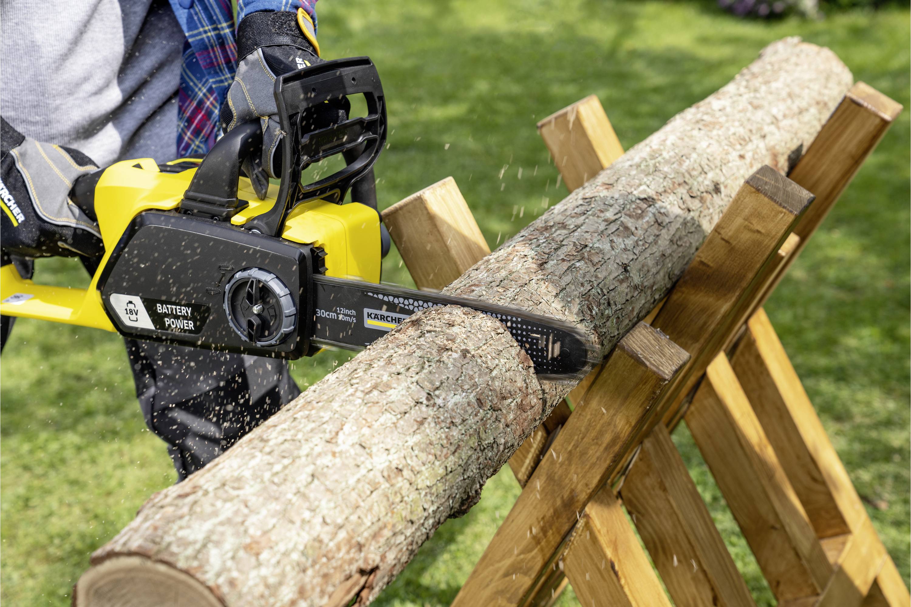 Person sägt mit einer elektrischen Kettensäge ein Holzstück, das auf einem Holzgestell liegt, im Garten.