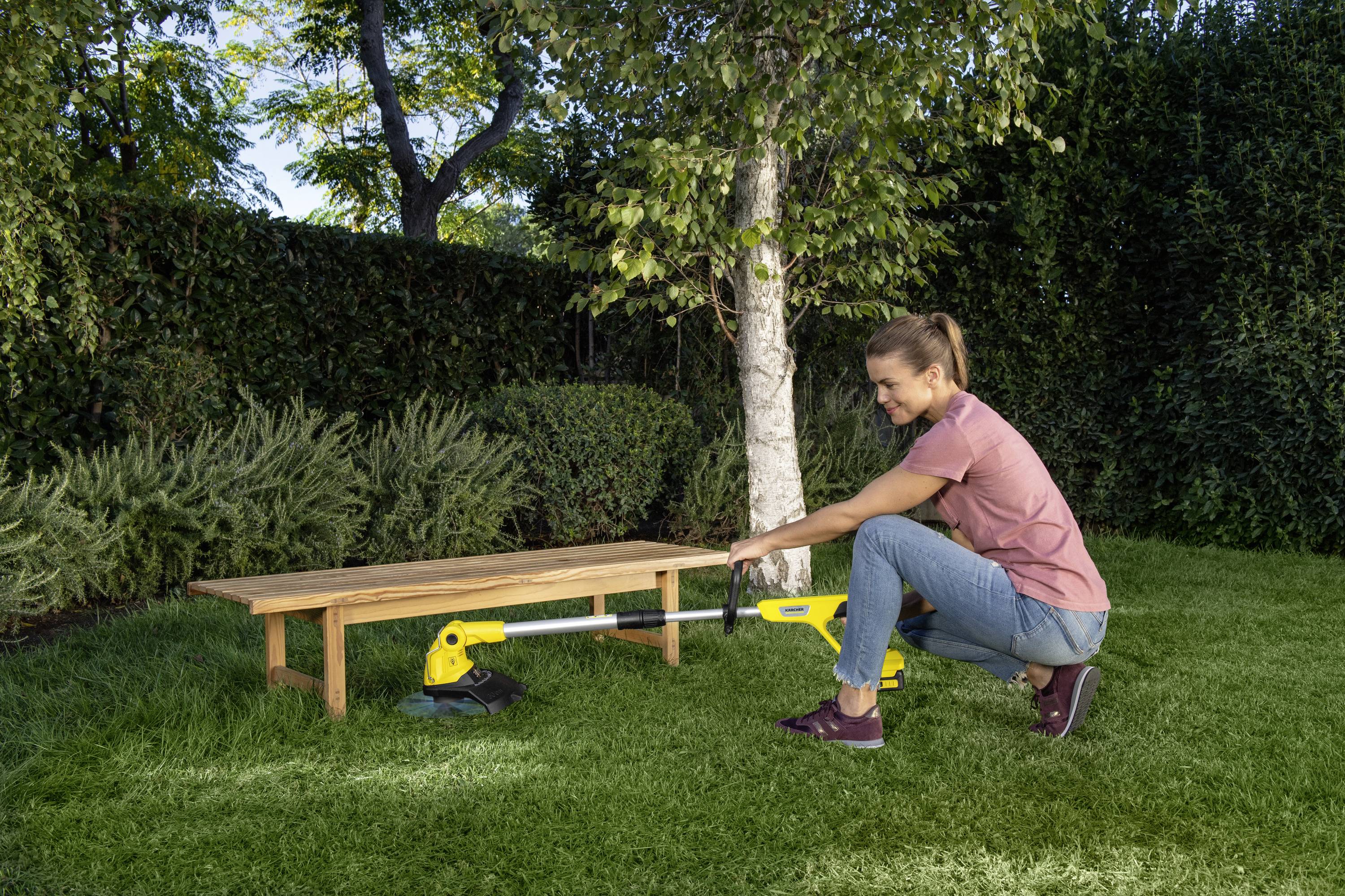 Eine Frau schneidet mit einem elektrischen Rasentrimmer das Gras in einem Garten. Im Hintergrund stehen Bäume und eine Holzbank.