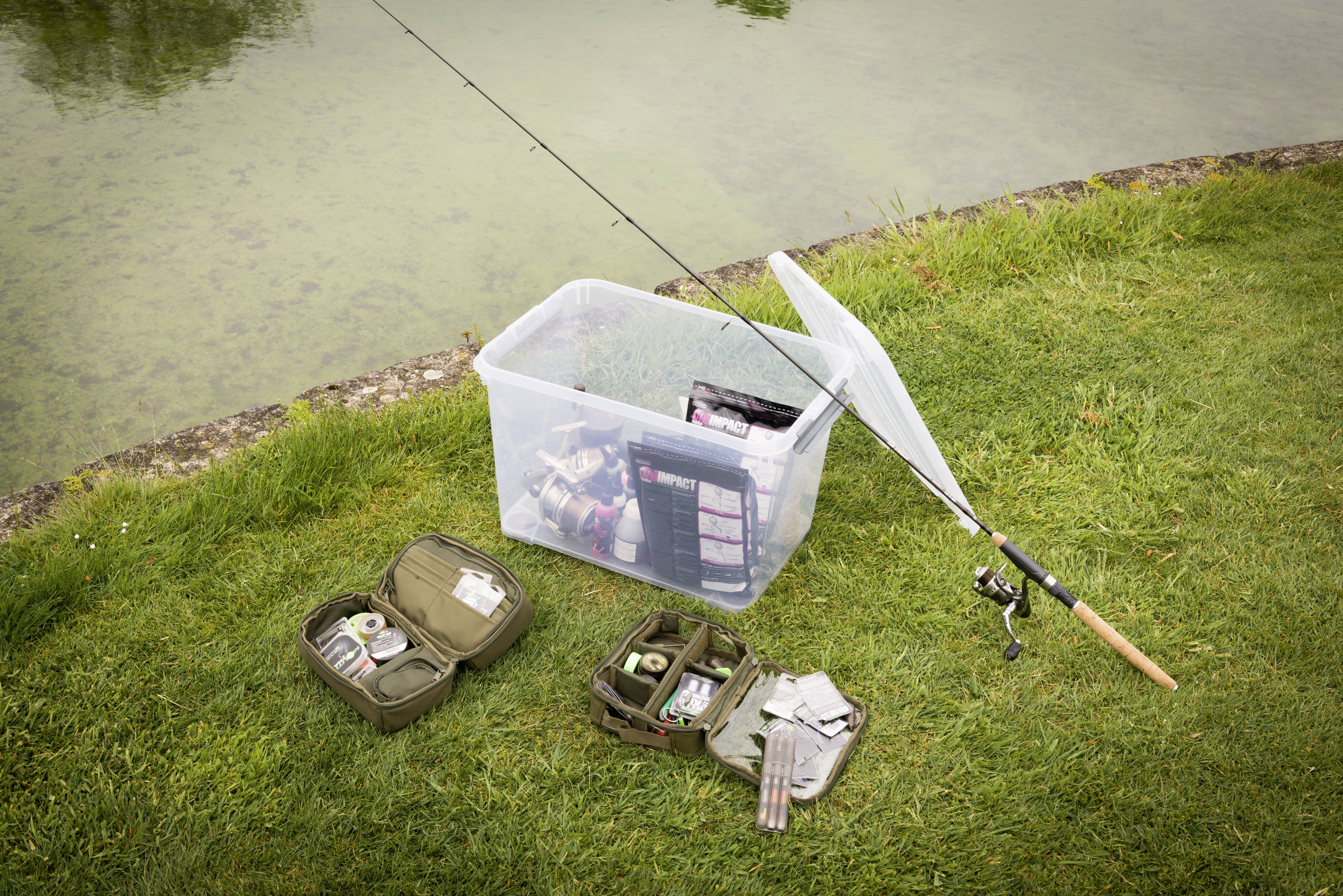Angelszene: Eine transparente Box mit Angelausrüstung steht neben einem Angelruten-Set am grasbewachsenen Ufer eines Sees.