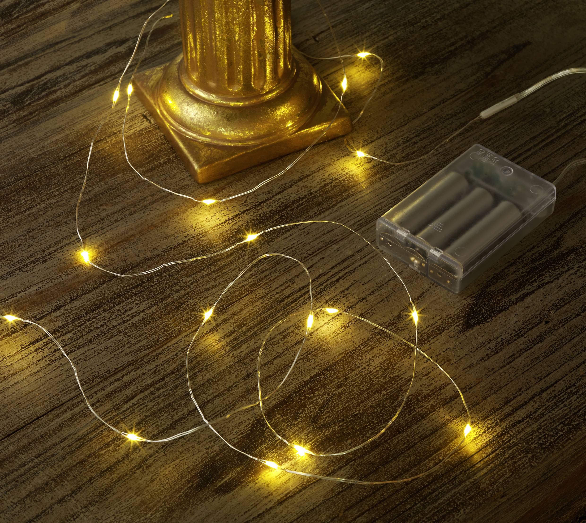 Lichterkette mit warmweißen LEDs auf Holztisch neben goldener Säule und transparentem Batteriefach.