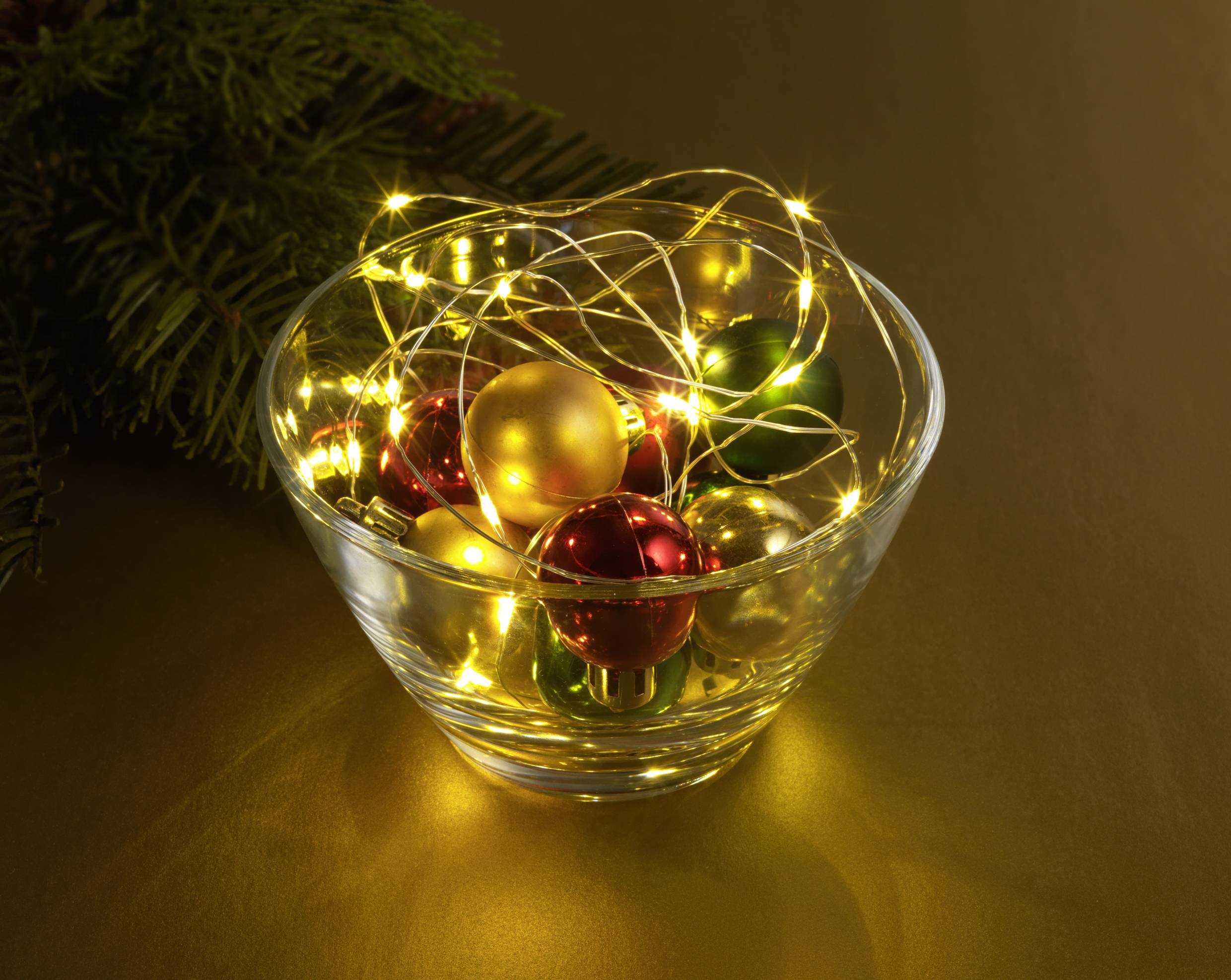 Eine Glasschale voller leuchtender Weihnachtskugeln und Lichterketten auf einem goldenen Hintergrund, daneben Tannenzweige.