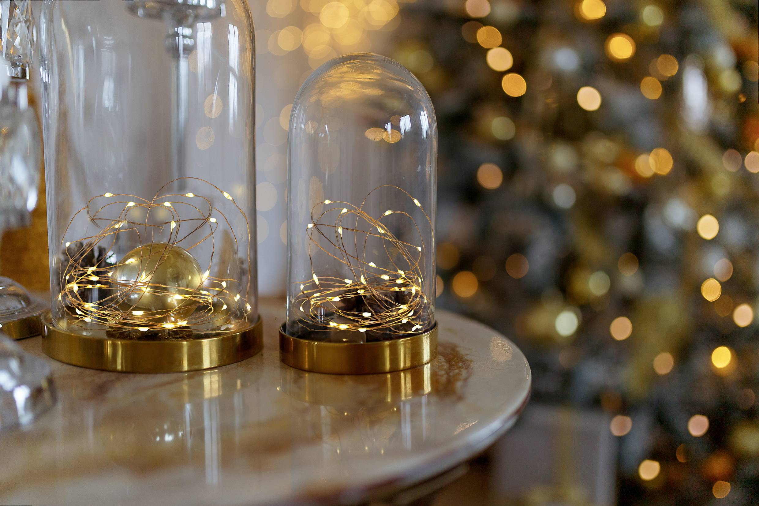 Glasglocken mit Lichterketten auf einem Tisch, im Hintergrund ein unscharfer, beleuchteter Weihnachtsbaum. Feiertagsstimmung und Dekoration.