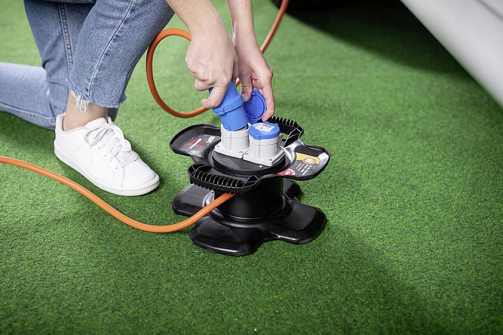 Eine Person steckt blaue Kabelstecker auf einem schwarzen Kabeltrommeladapter ein, der auf grünem Kunstrasen liegt.