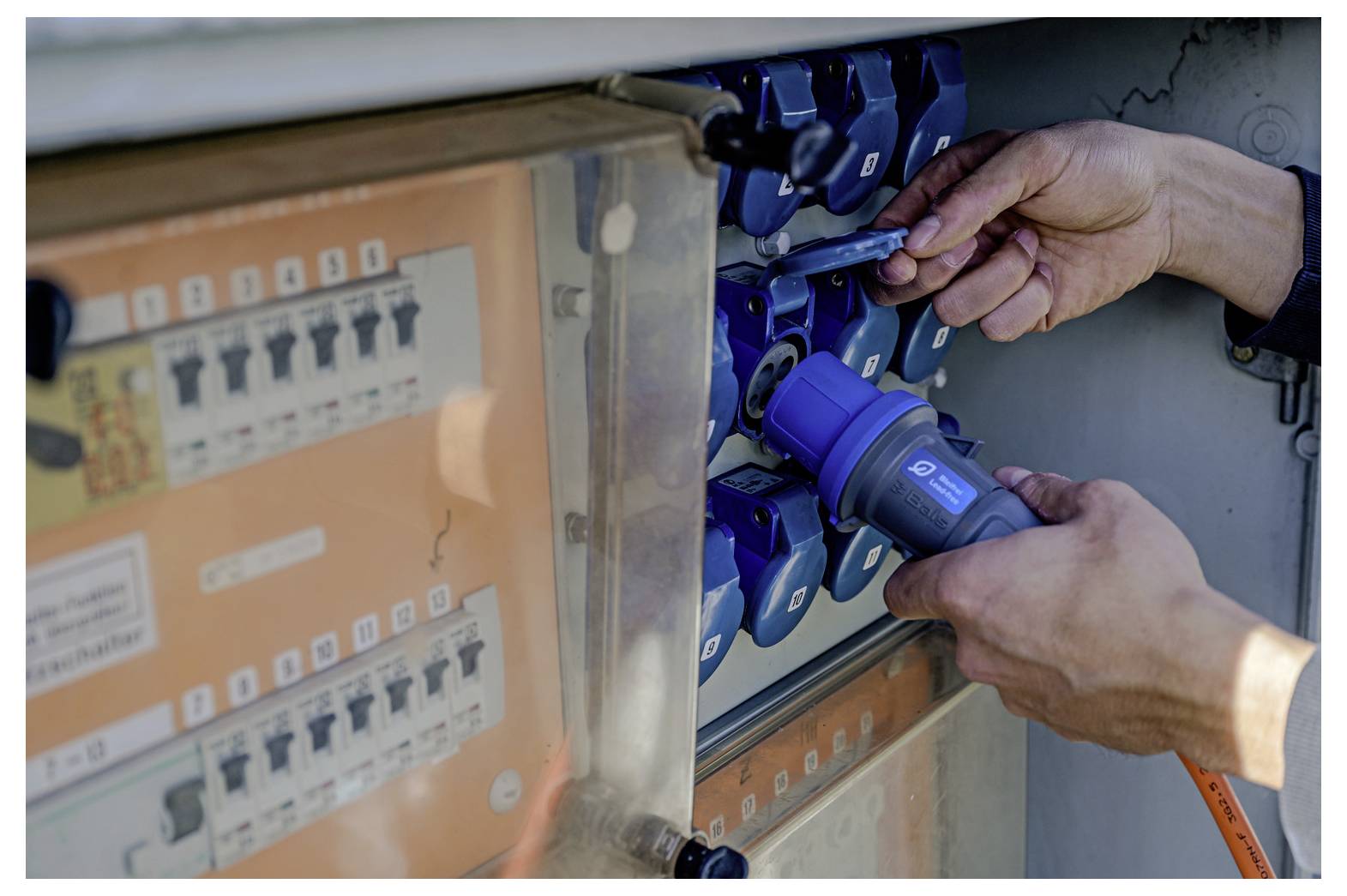 Hände, die blaue Elektrostecker in ein Stromverteilerpanel mit beschrifteten Schaltern stecken.