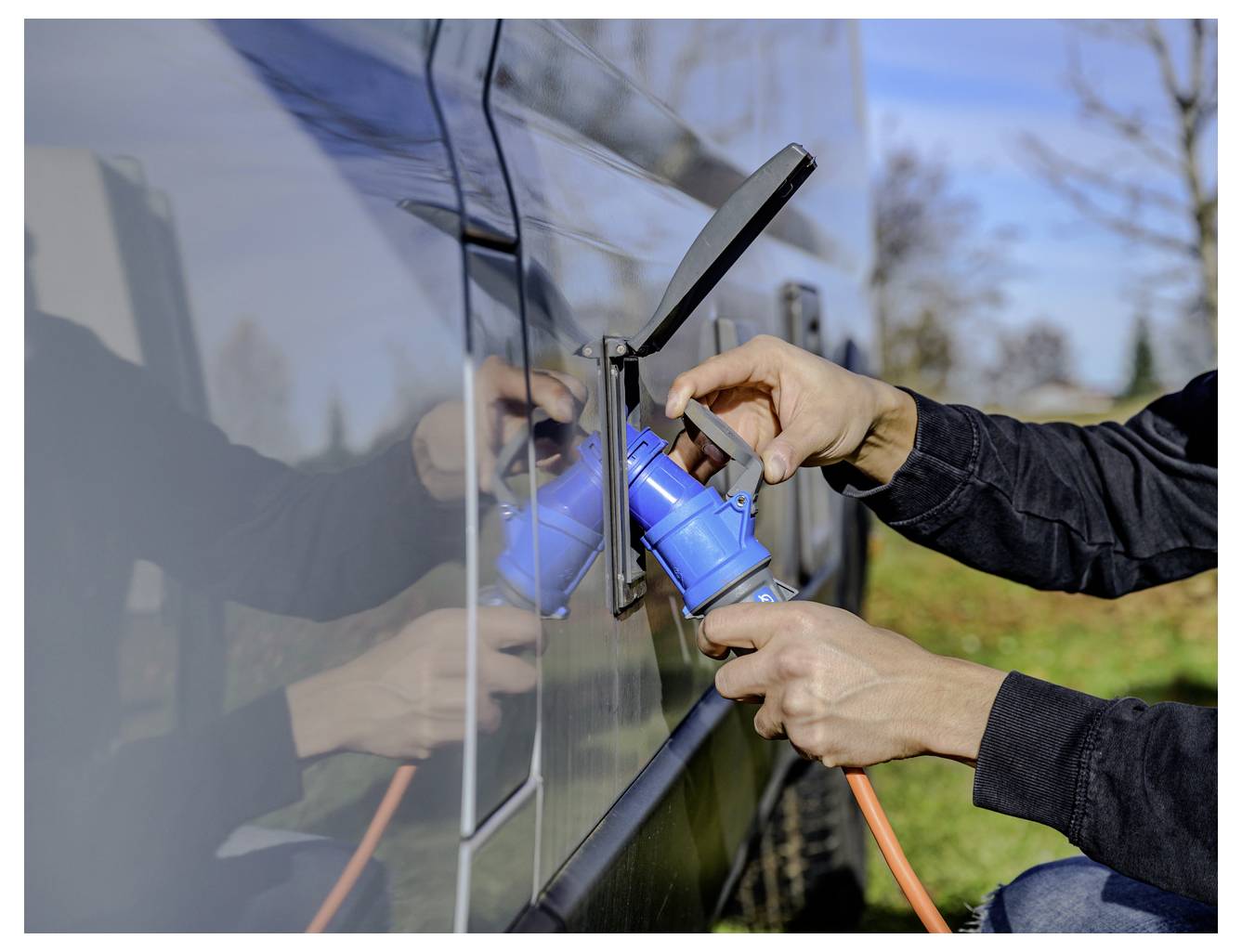 Eine Person steckt ein oranges Kabel in eine blaue Ladebuchse an einem Fahrzeug und deutet auf das Laden eines Elektrofahrzeugs hin.